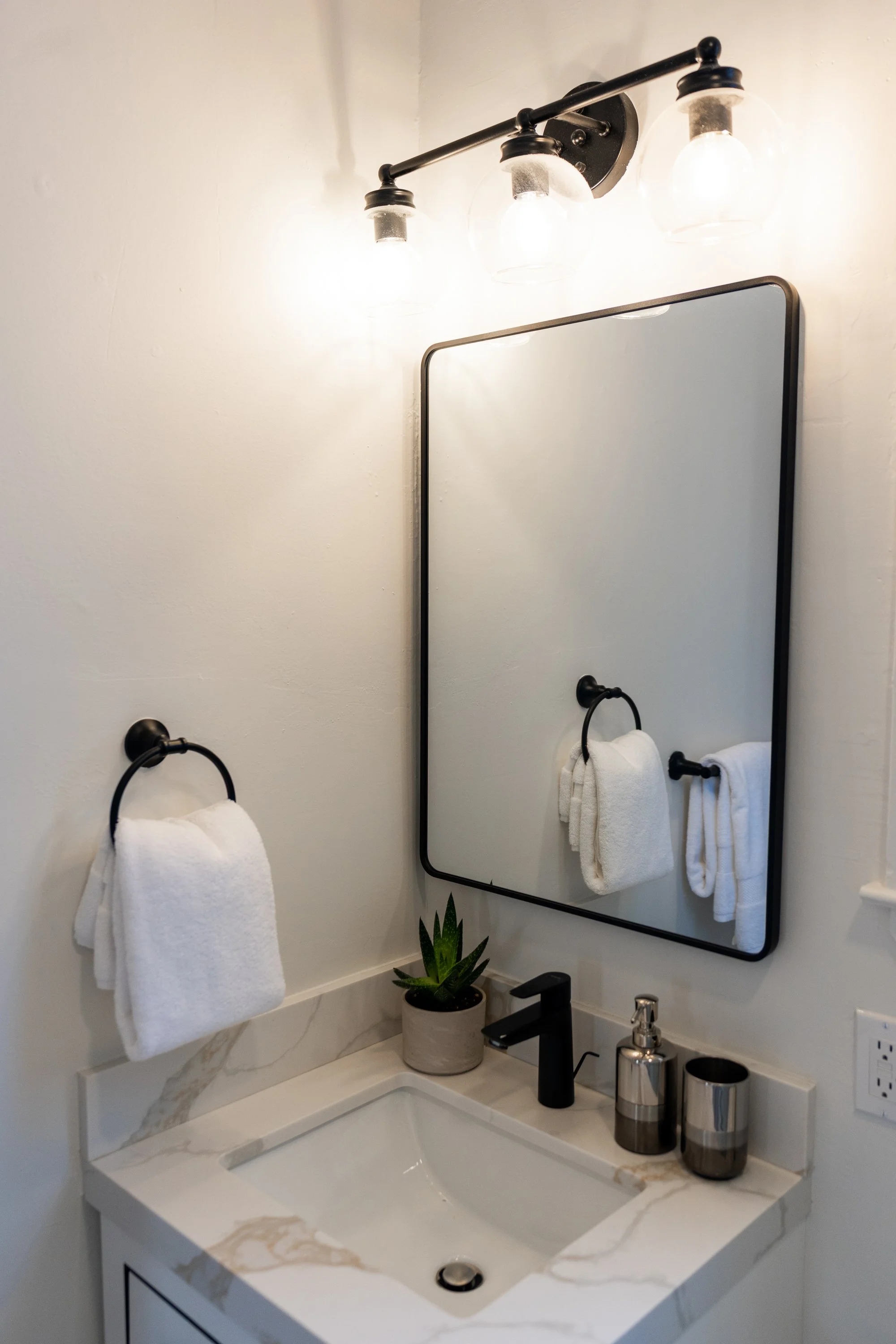 Bathroom sink with black faucet, two black and silver soap dispensers, a potted plant, towel rings with white towels, mirror, and modern light fixture with four glass bulbs.