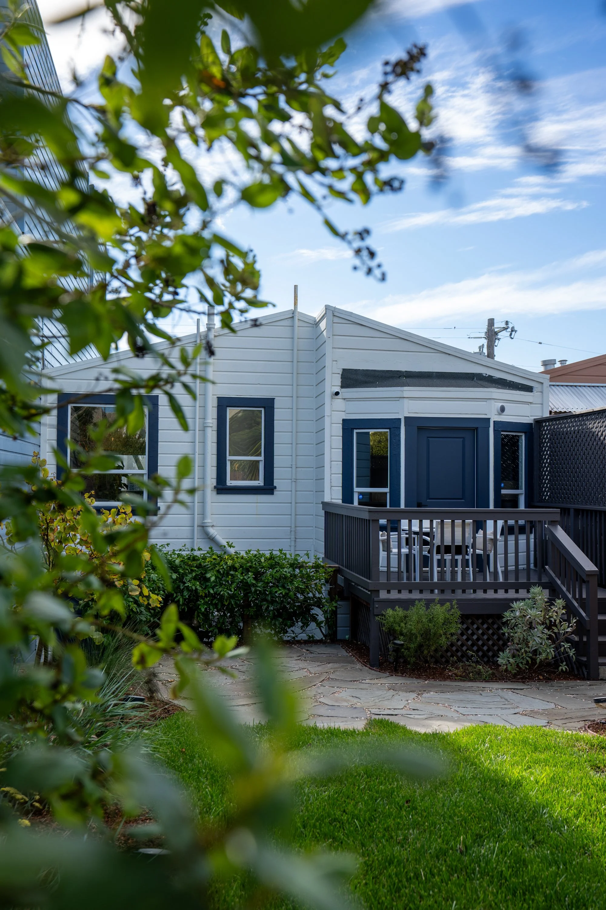 A backyard with a lush green lawn, potted plants, and a gray wooden deck with a black railing, leading to a white house with blue window frames. The house has a sloped roof, drainage pipes, and a window with curtains. There are trees and a power pole