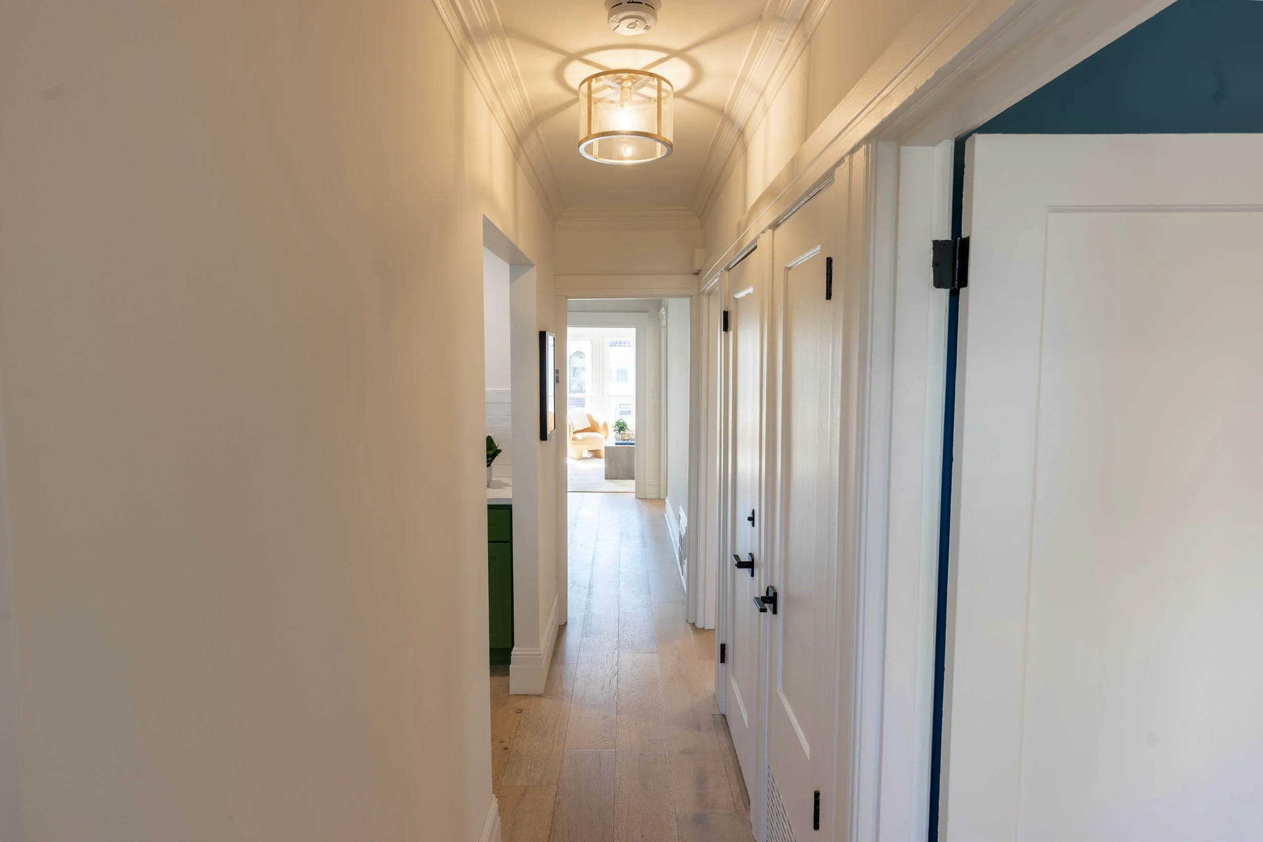A bright hallway with beige walls, wooden flooring, white crown molding, and a ceiling light fixture. At the end of the hallway, there is a sitting area with a yellow armchair, a window, and a small table with a plant.