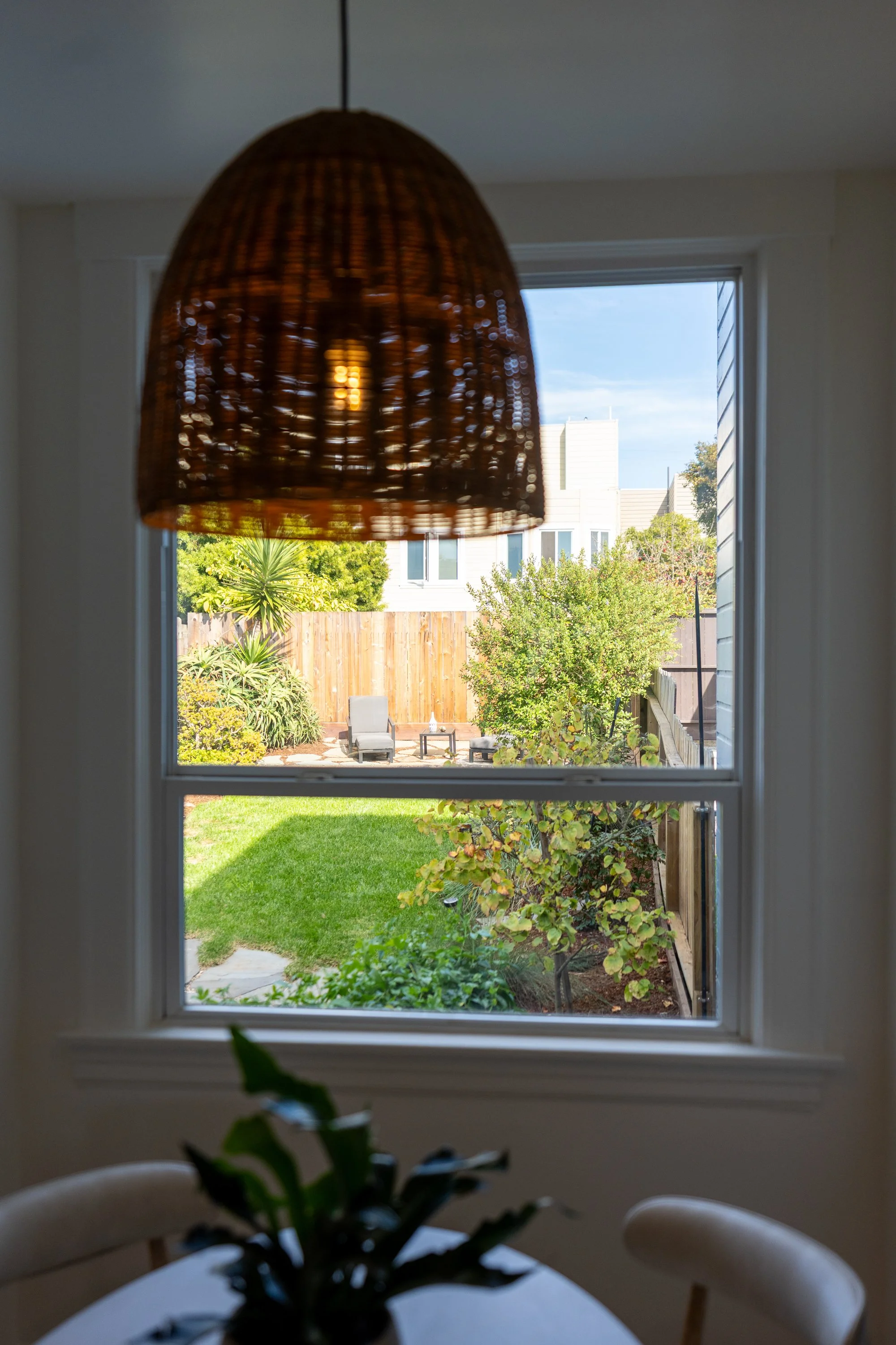 View of a backyard garden through a window with a wicker lampshade hanging from the ceiling in the foreground.