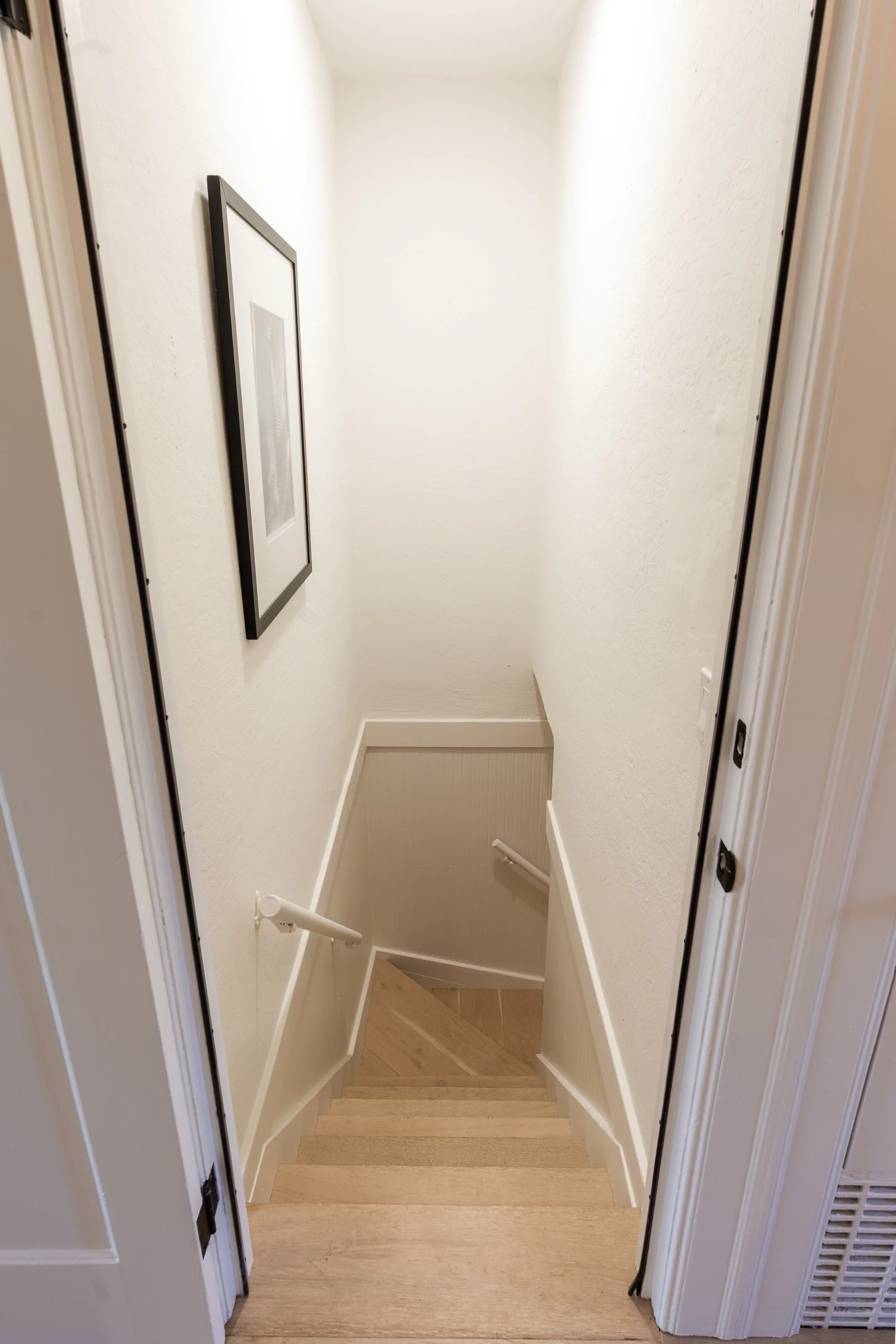 A staircase in a home with white walls, a handrail, and a framed picture on the left wall, leading down to a lower level with beige flooring.