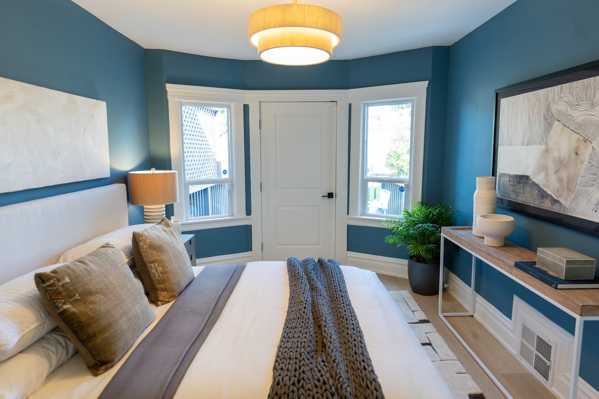 A bedroom with blue walls, a white bed with beige and brown pillows, a wooden nightstand with a beige lamp, a large framed black-and-white abstract artwork, a small white table with decorative vases, two windows, and a potted plant near the windows.