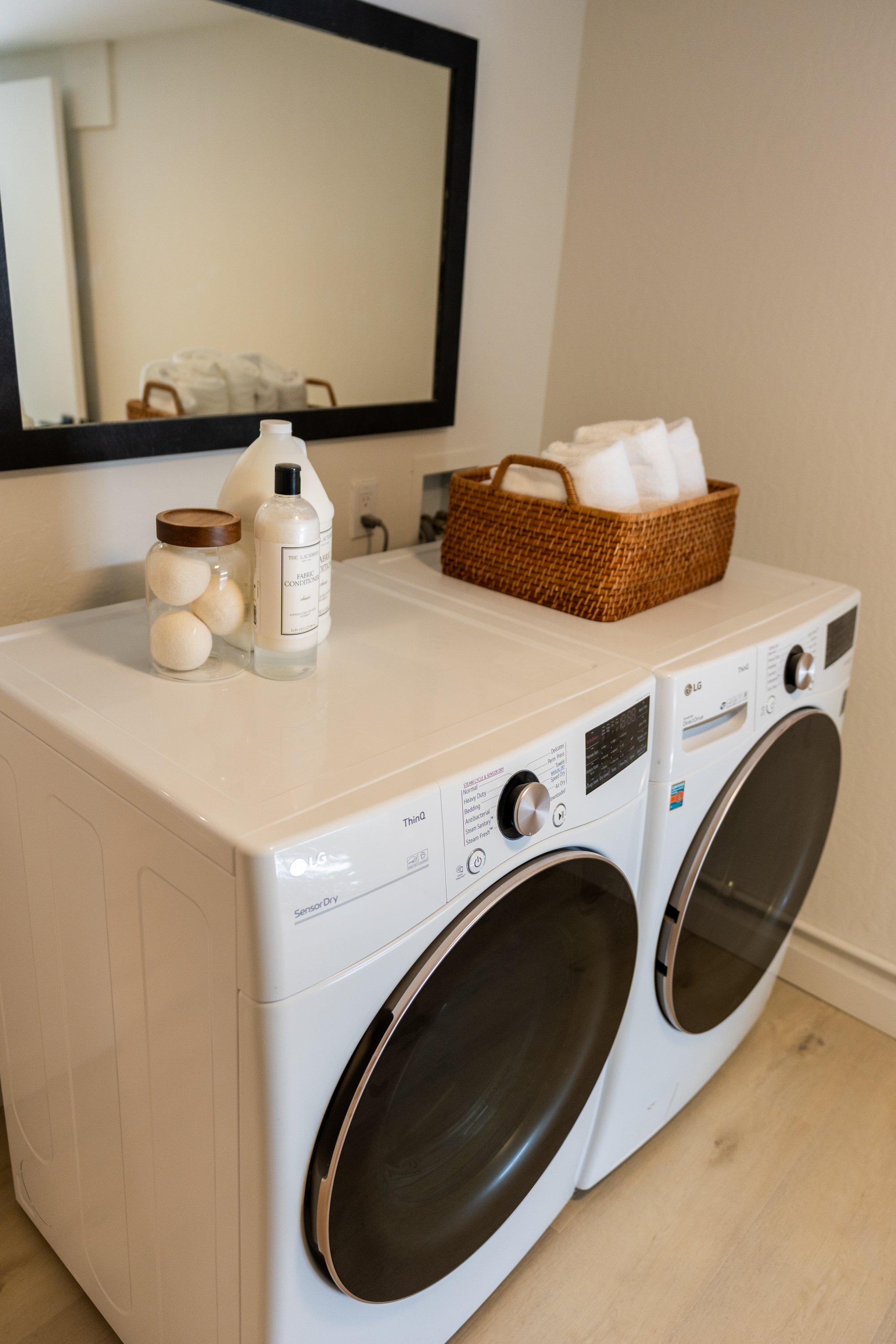 A laundry room with a white washer and dryer side by side. On top of the washer, there is a glass jar with white laundry balls, a white bottle, and a smaller bottle of fabric conditioner. A brown wicker basket holding rolled white towels is on top of