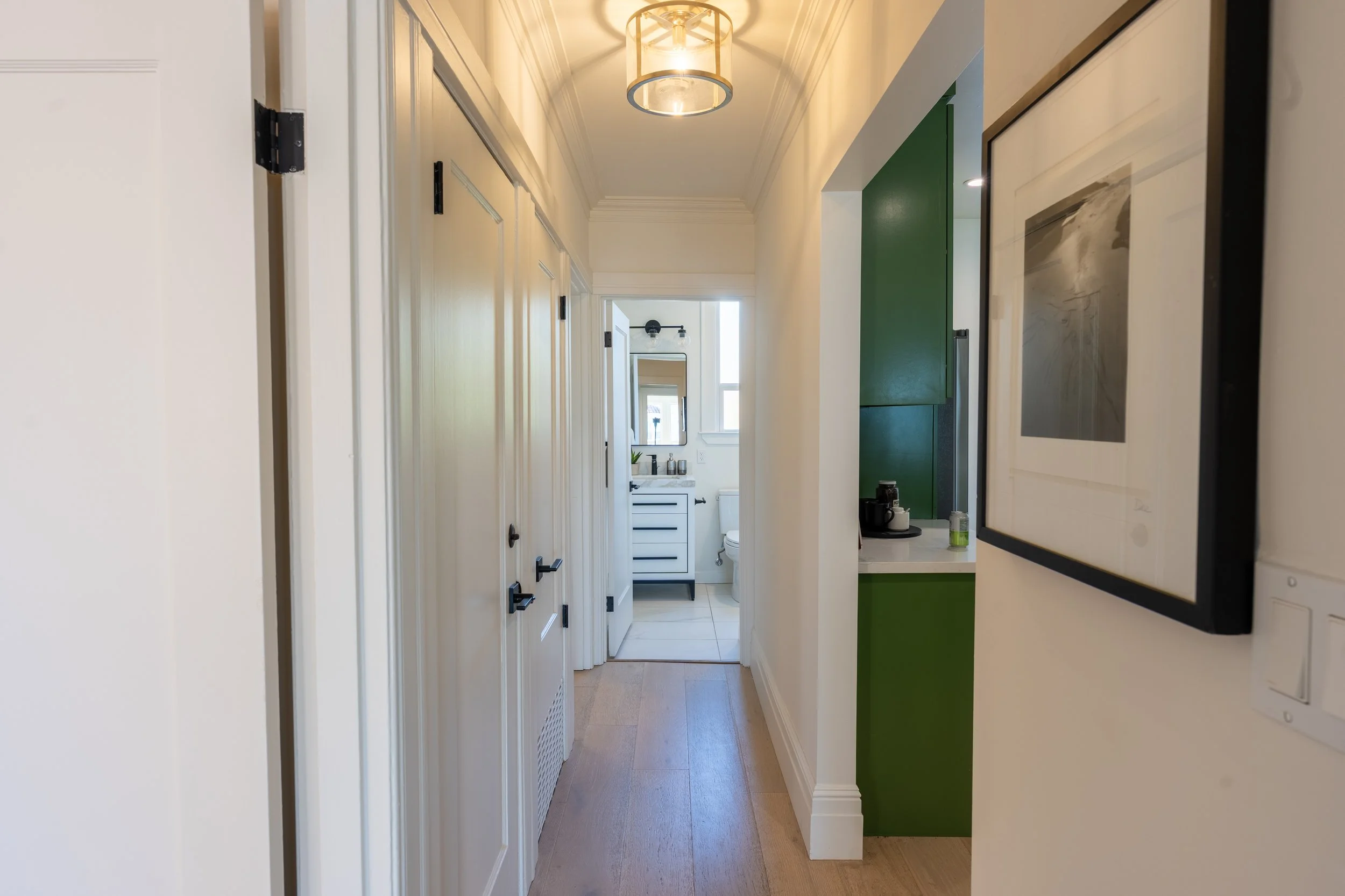 A narrow hallway leading to a bathroom, with white walls, wooden floors, a green accent wall, framed artwork, and light fixtures.