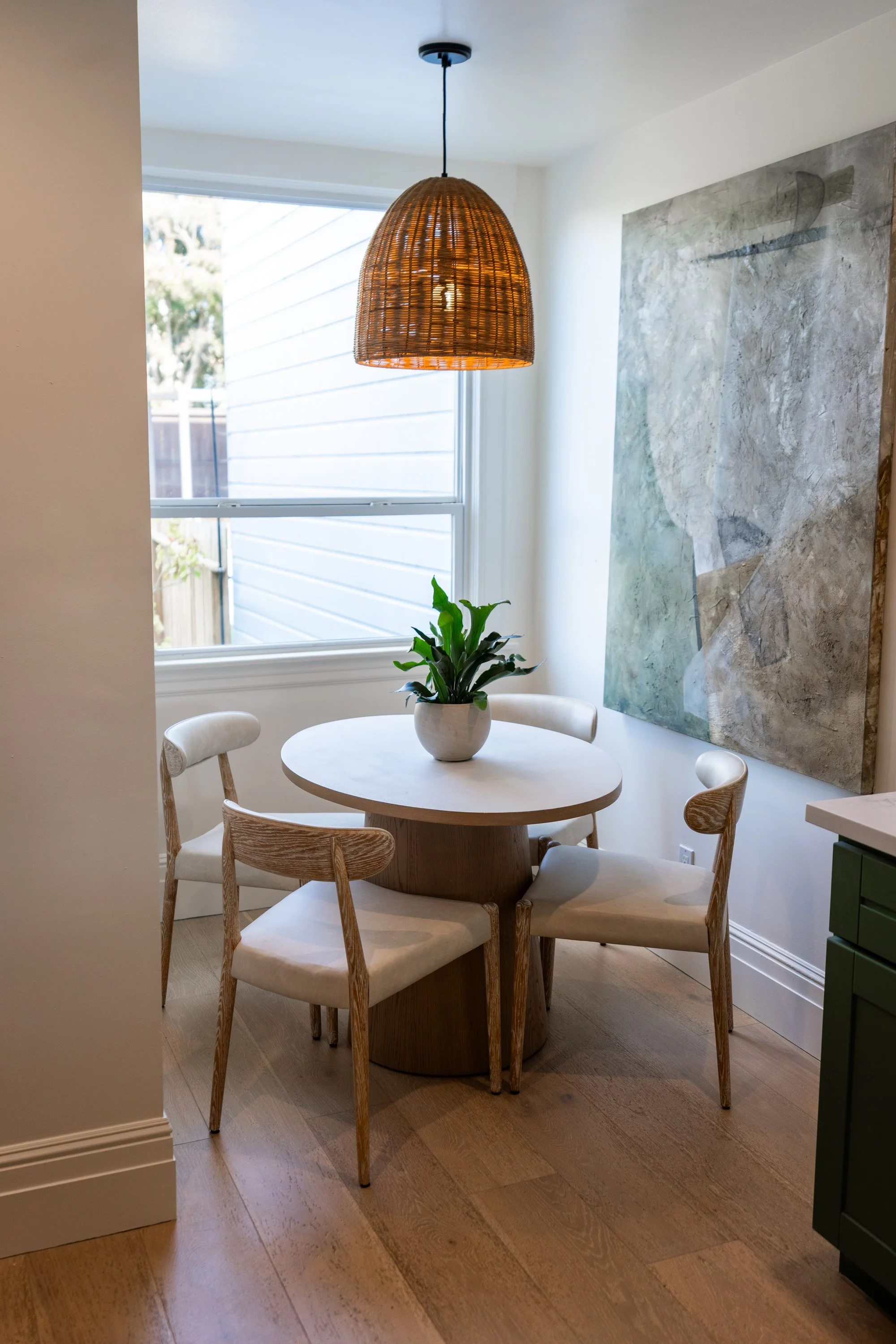 A round white dining table with four wooden chairs, a potted plant in a white pot on the table, a large abstract painting on the wall, a wooden pendant light hanging from the ceiling, a window with outside view, and a green cabinet.