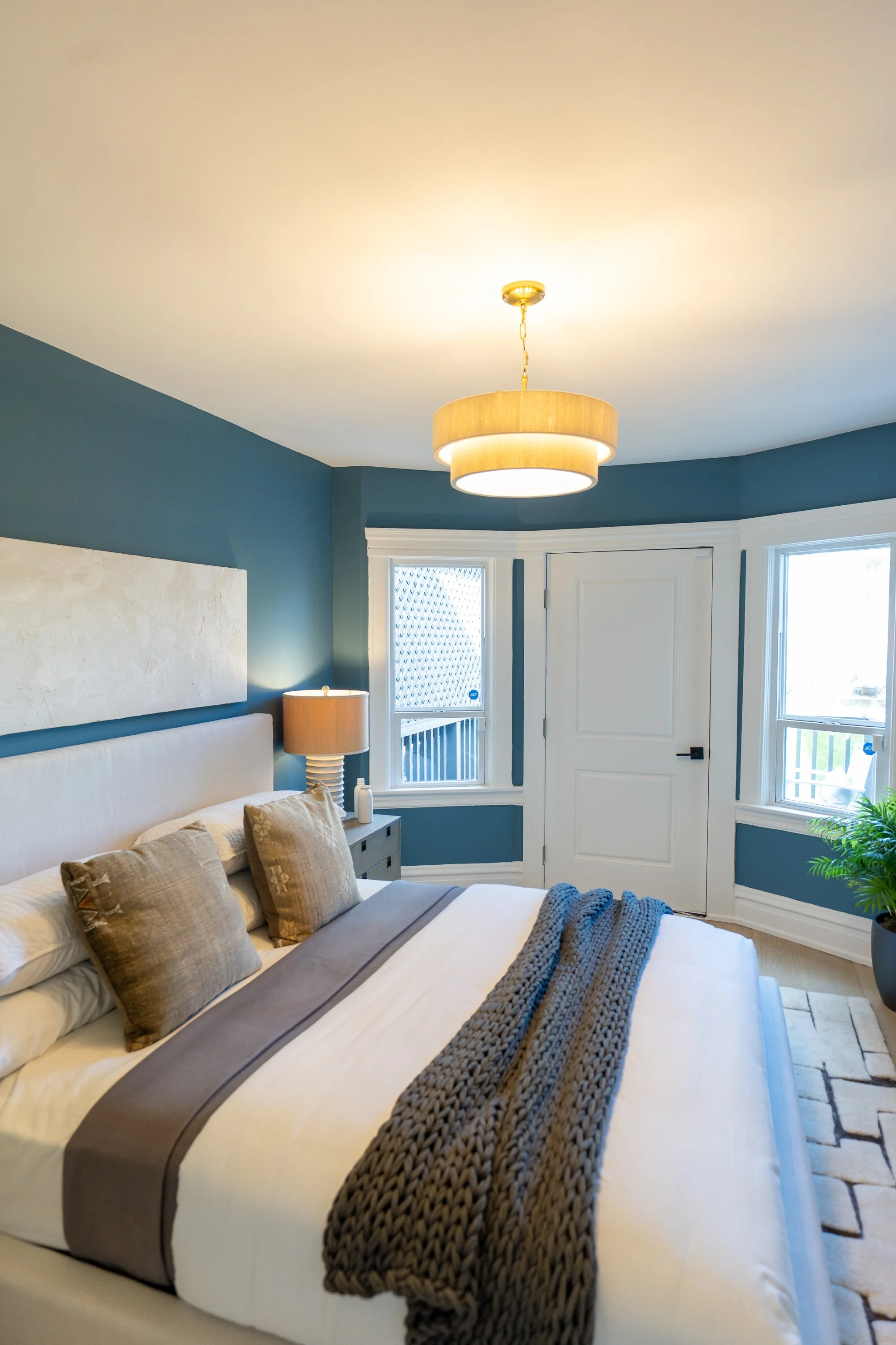 A bedroom with a large bed, multiple pillows, a side table with a lamp, and a patterned blanket. The walls are painted blue, with windows letting in natural light. A ceiling light fixture is also visible.
