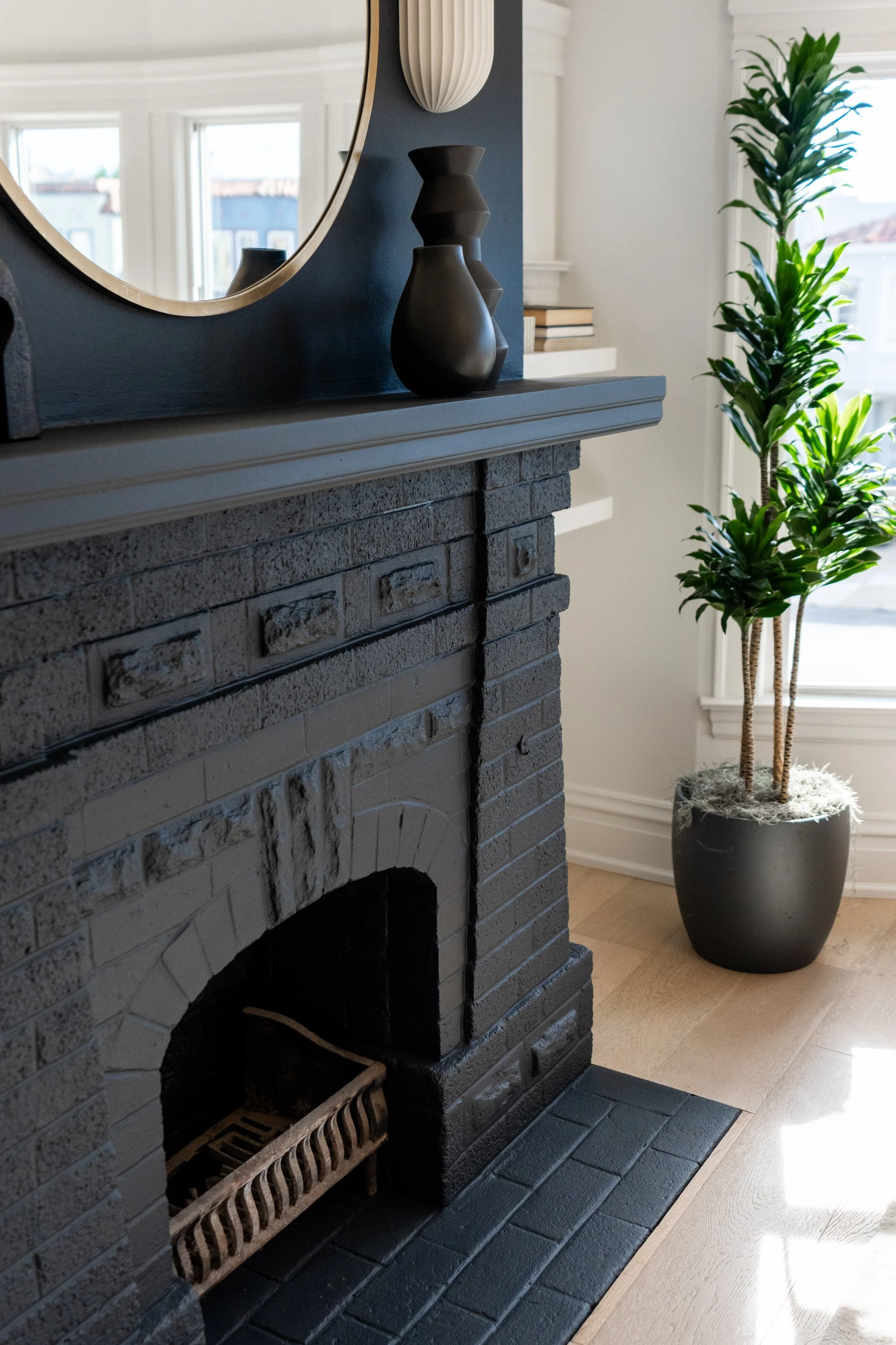 Close-up of a black brick fireplace mantel decorated with black and beige vases, with a mirror partly visible above. To the right, a tall potted plant with green leaves is positioned near a window, allowing sunlight to illuminate the scene.