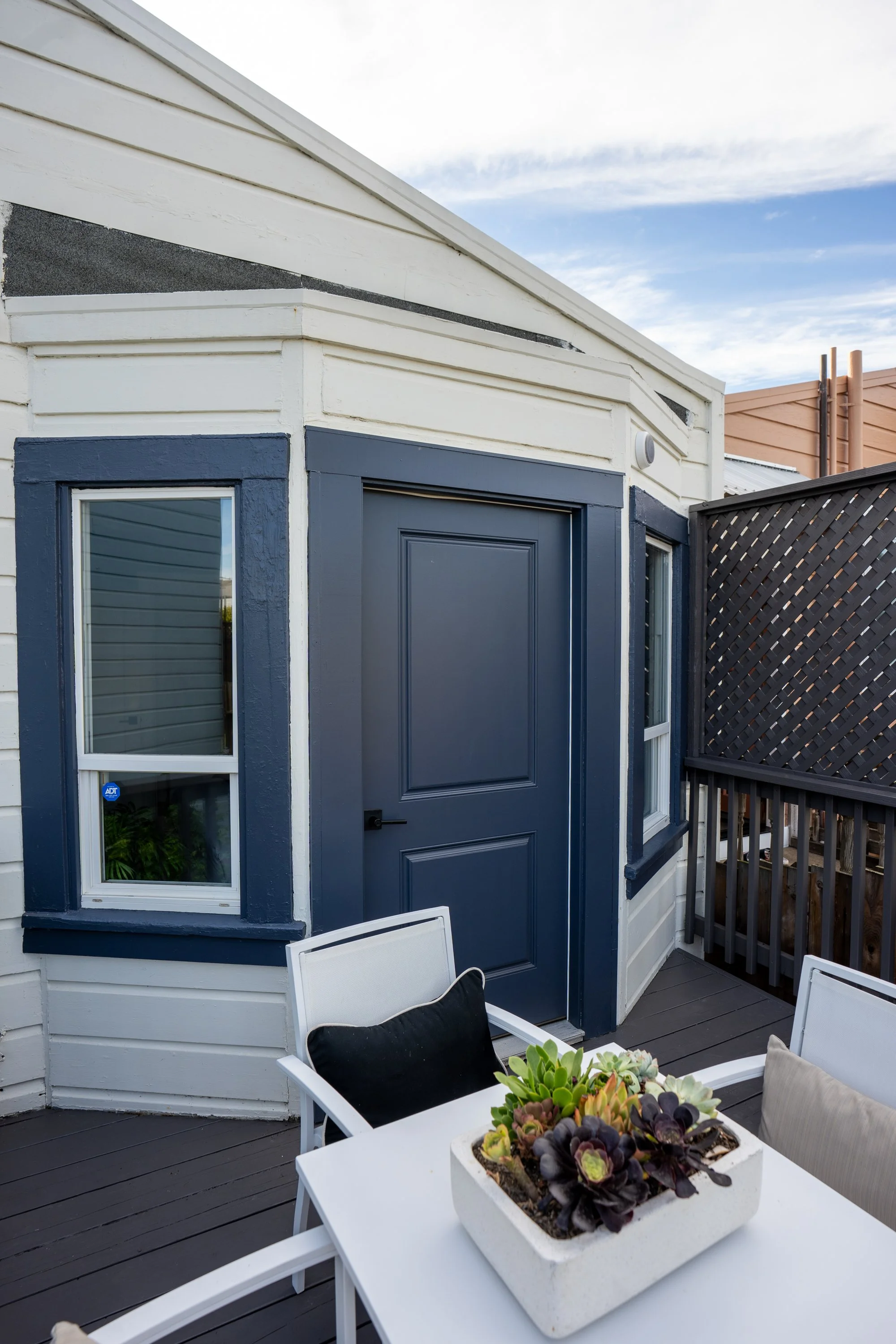 A rooftop patio with a white table and chairs featuring a rectangular planter with succulents, a blue door, two windows, gray floorboards, a dark wood lattice fence, and a partly cloudy sky.