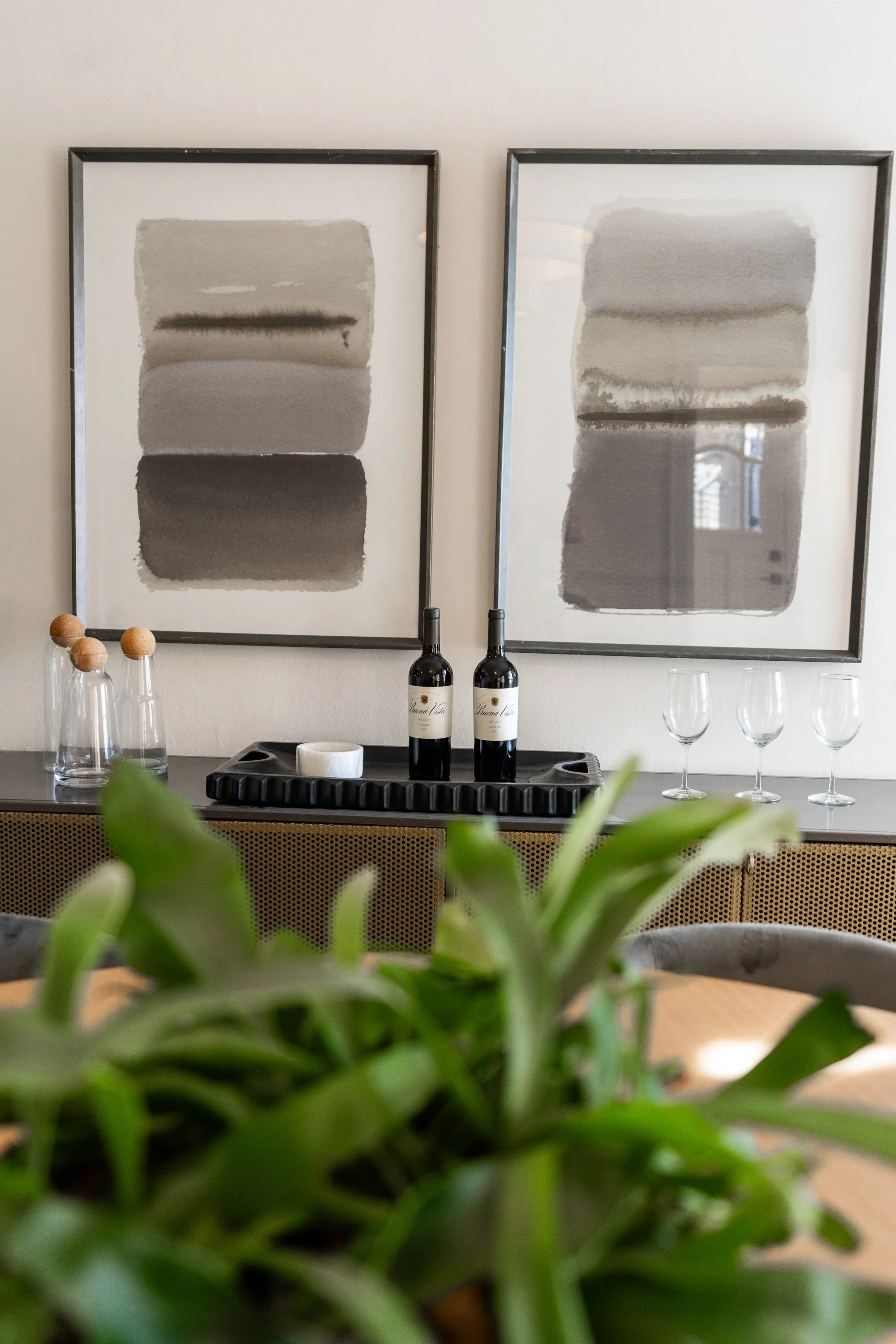 Two framed abstract black, gray, and white watercolor paintings hanging on a white wall. Below the paintings, two bottles of wine, a marble container, and several wine glasses are displayed on a black tray. Green plants are slightly visible in the fo