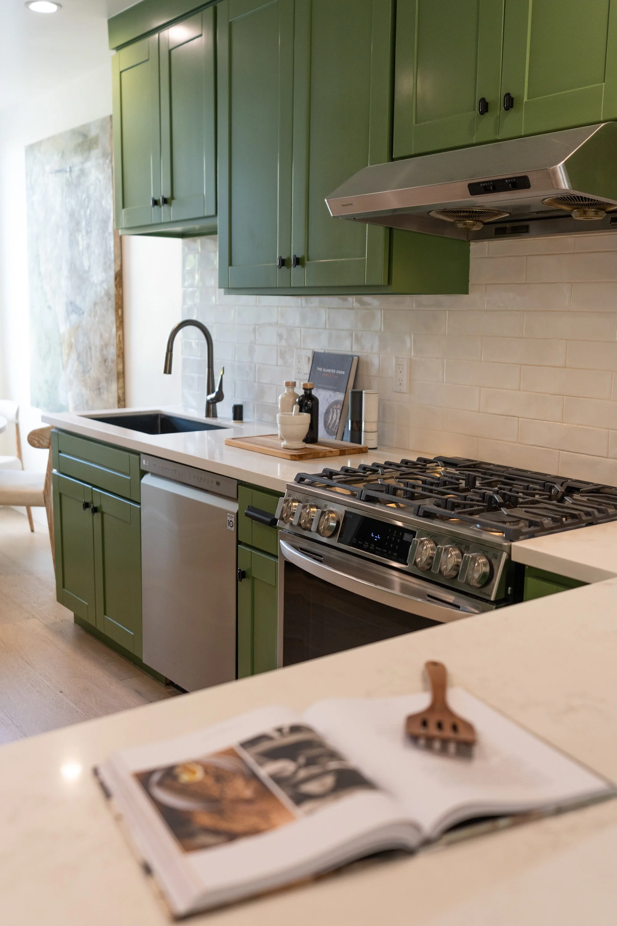 Modern kitchen with green cabinets, a white countertop, a black sink, and a stainless steel stove and range hood. There are small kitchen items and a framed book on the counter.