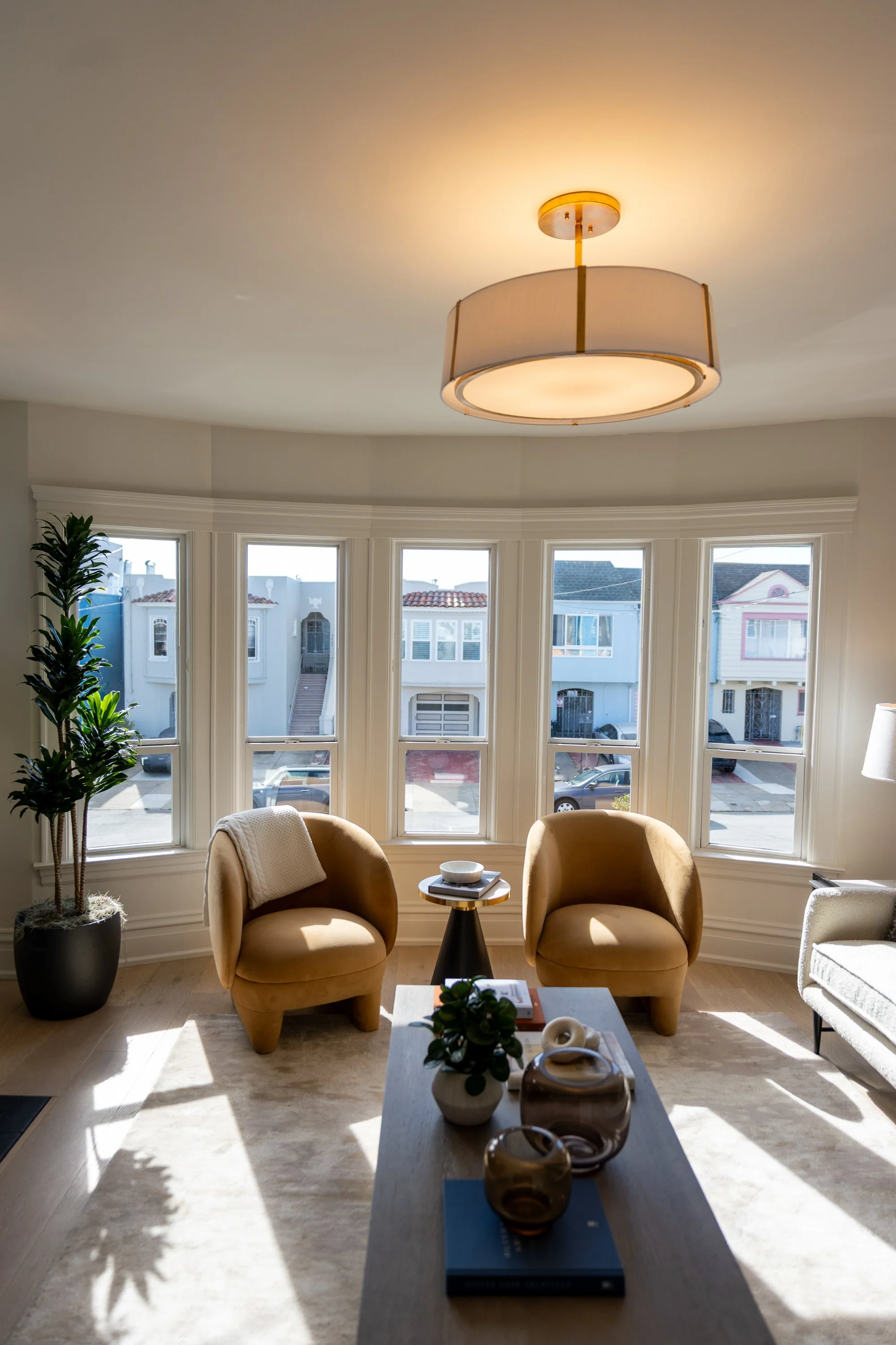 Living room with a large bay window, two tan armchairs, a small table, indoor plants, and a ceiling light fixture. Sunlight streams through the windows, illuminating the room.