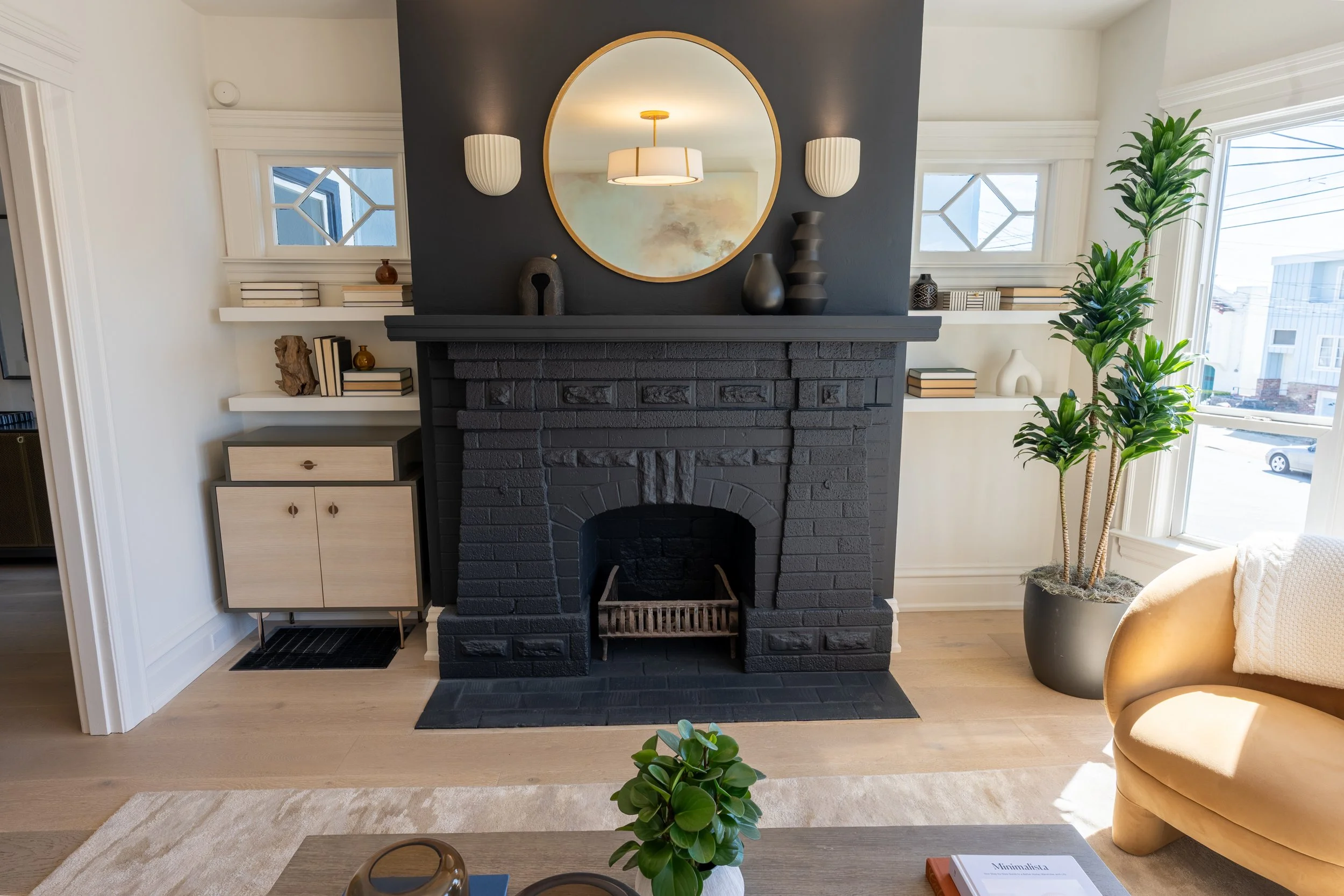 Living room with a black brick fireplace, decorative vases, circular mirror above, two white wall sconces, two windows with diamond-shaped panes, a large potted plant, a beige armchair, and built-in shelves with books and decor.