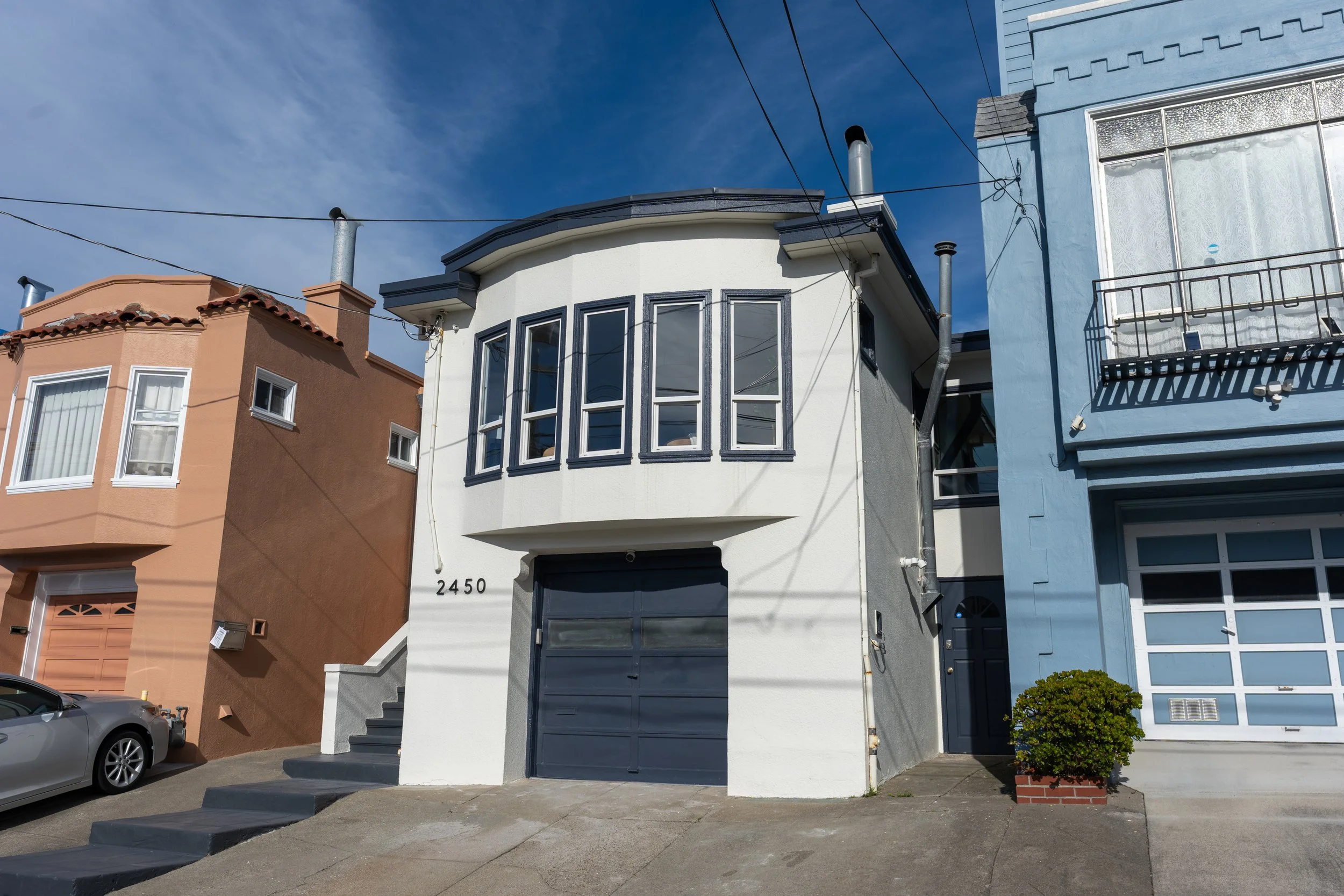 A row of colorful houses on a sunny day, with a gray car parked in front of a white house. The houses are painted in pastel shades including peach, white, and blue, with distinct architectural details. The white house has a curved front with black-fr