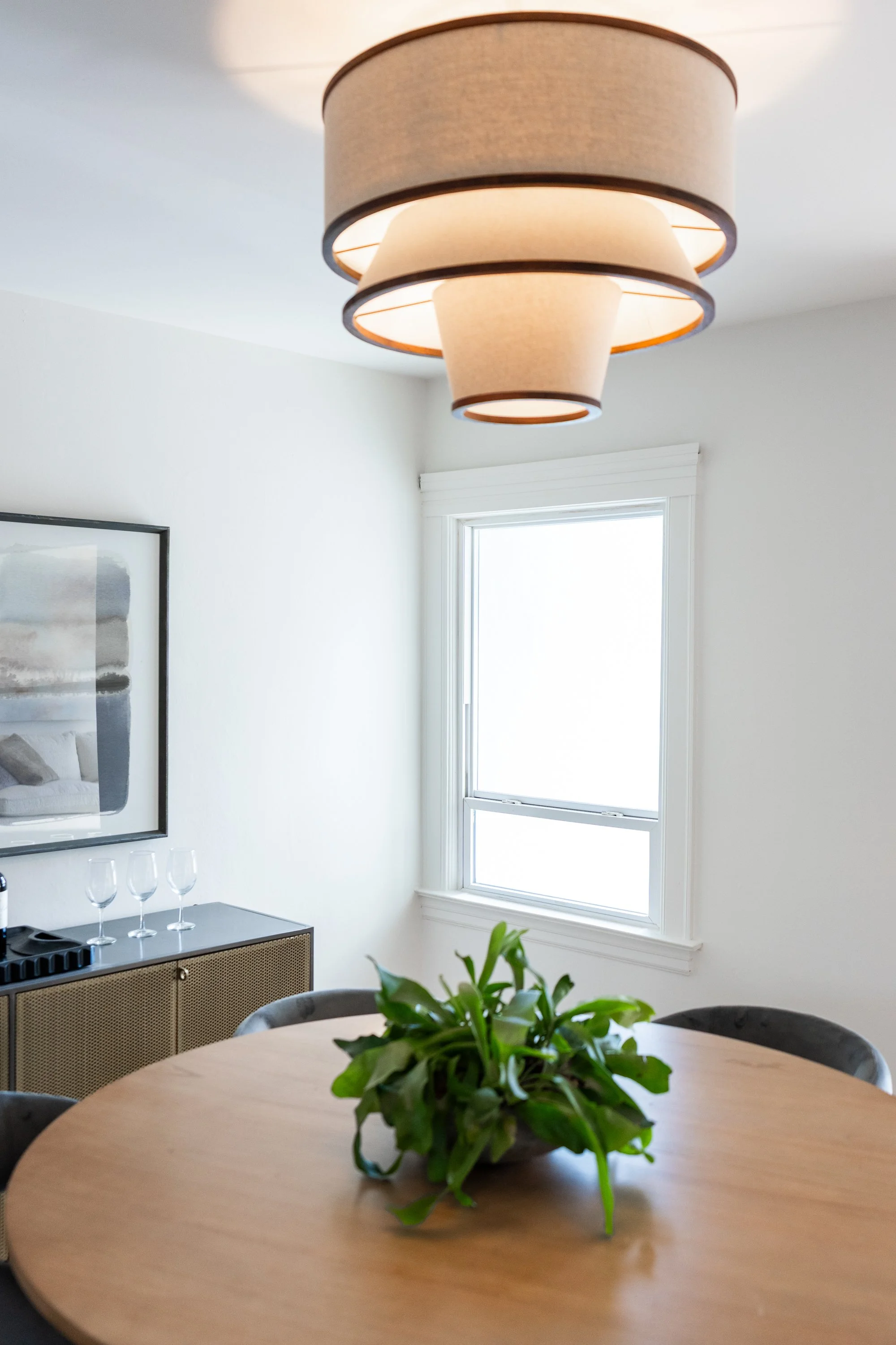 A modern living room with a round wooden table, a green potted plant in the center, a large window allowing natural light, a beige and black overhead light fixture, wall art, and a black sideboard with glassware.