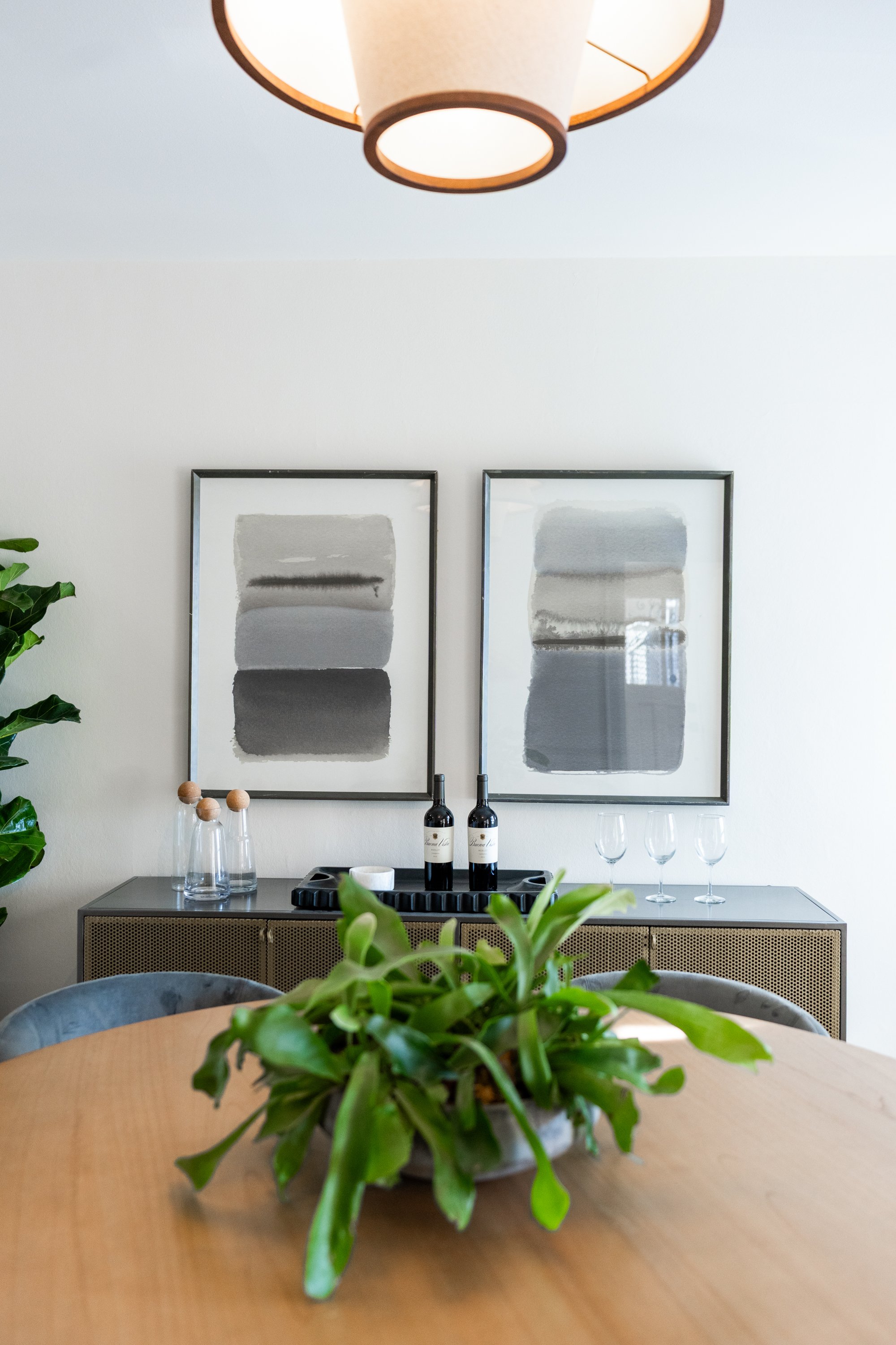 A dining room table with a green potted plant in the foreground, a sideboard with two bottles of wine, glasses, and decorative objects, and three framed abstract art pieces on the wall, with a hanging ceiling light above.
