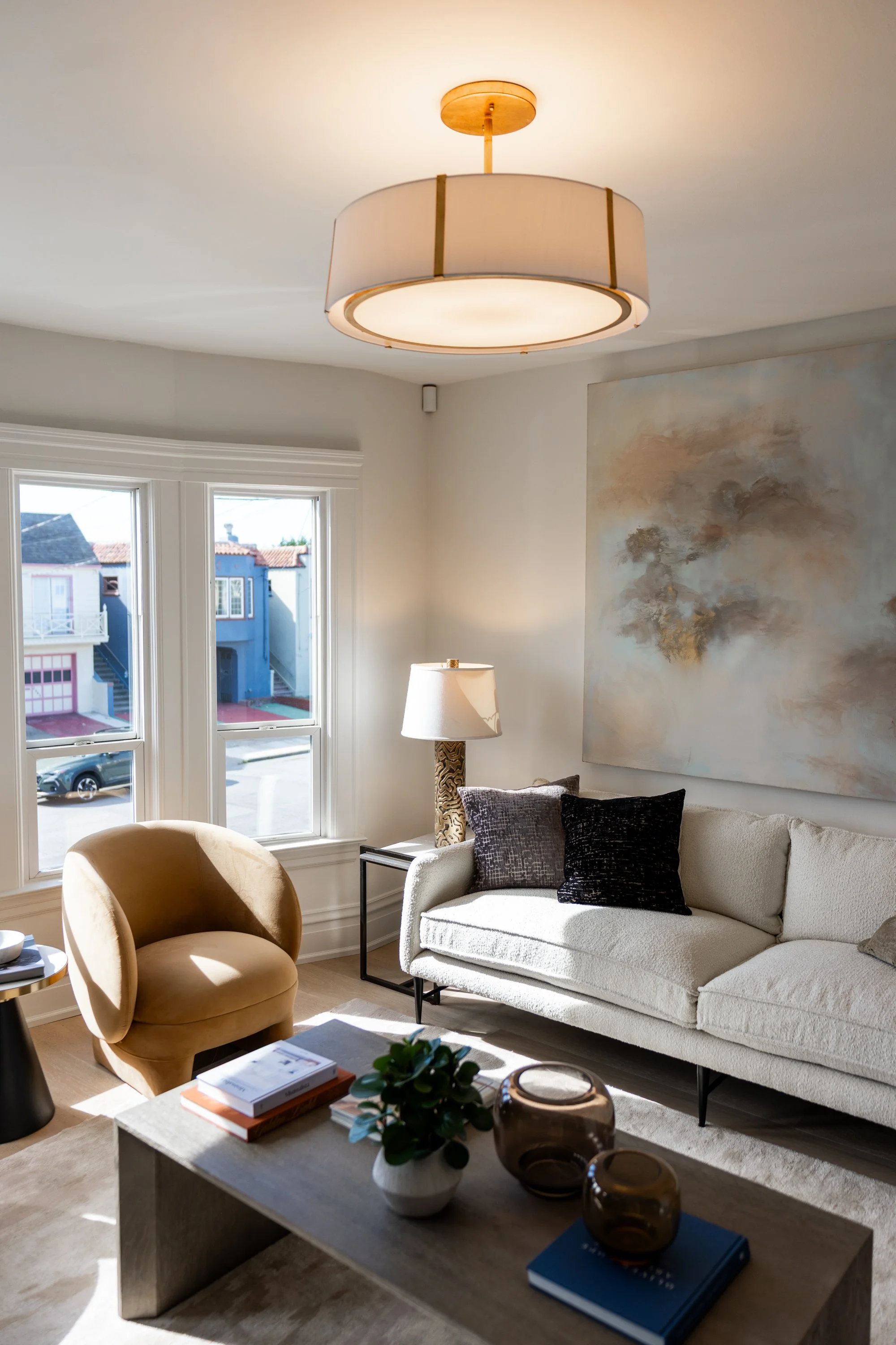 Living room with a round ceiling light, a white sofa with dark pillows, a beige armchair, a table with books and decorative items, large windows showing colorful buildings outside, and a large abstract wall art.