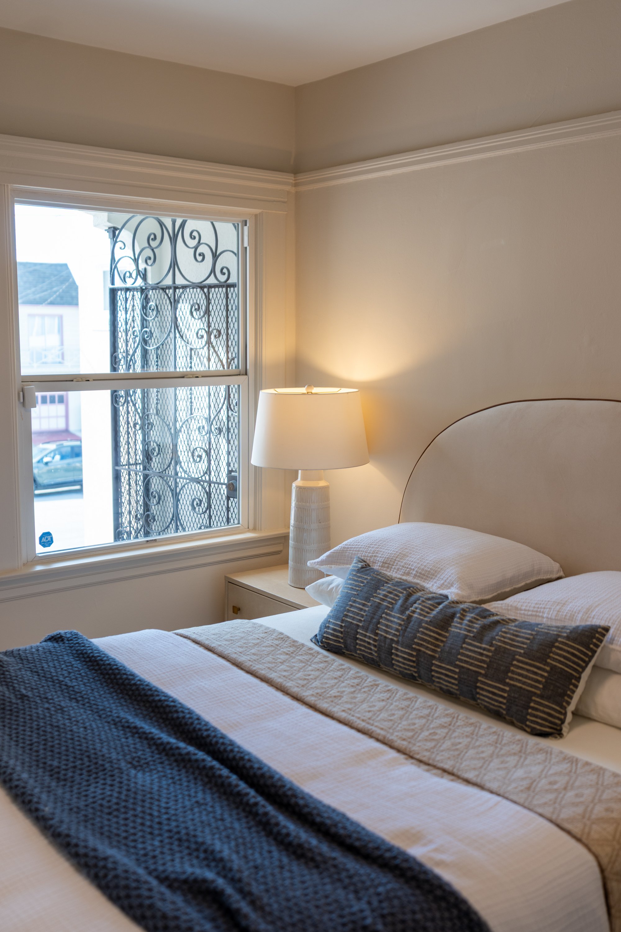 A bedroom with a window showing decorative metal bars and a street outside, a bed with a beige headboard, white bedding, a striped pillow, a beige quilt, and a dark blue throw blanket. A white table lamp with a textured base is on a nightstand beside