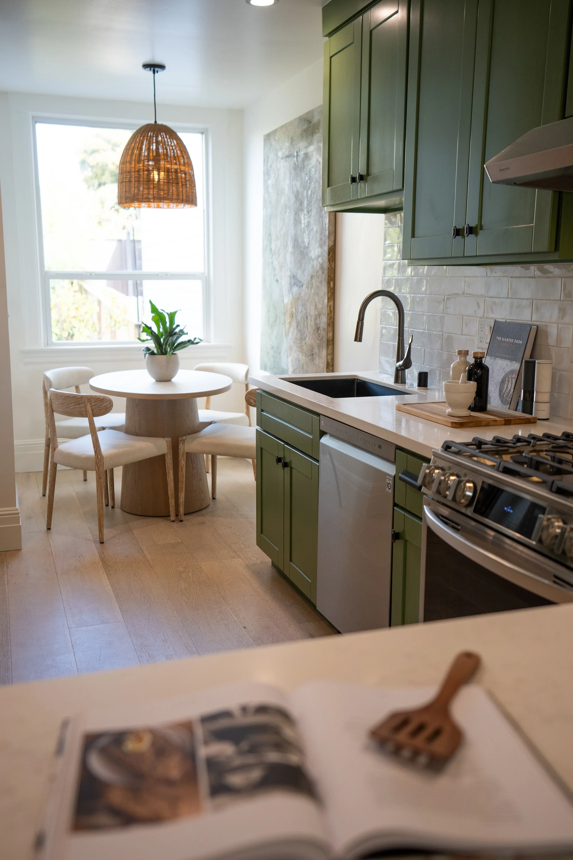 A kitchen with green cabinets, a black sink, a marble countertop, a white tile backsplash, and a window with a woven pendant light hanging above a round dining table with white chairs. There are decorative bottles and a book on the counter. A magazin