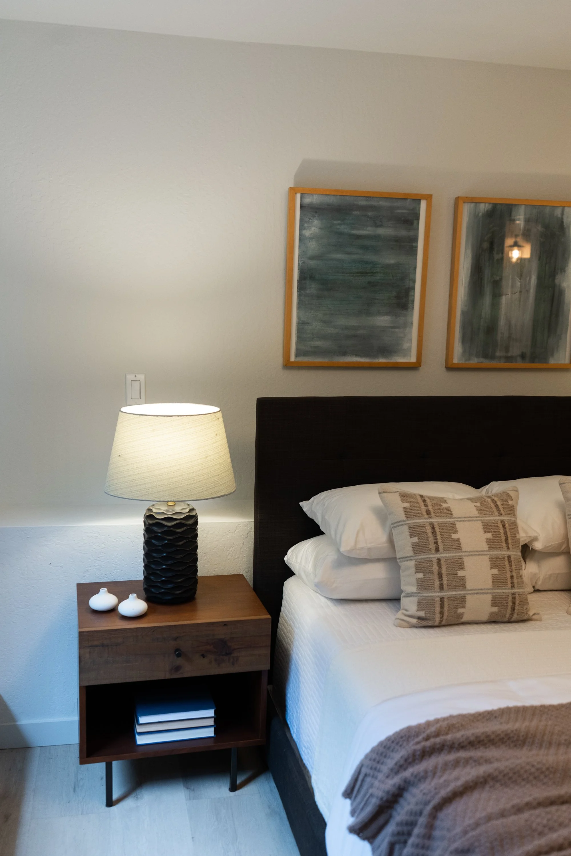 A bedroom with a bed, wooden nightstand, lamp, pillows, and wall decor.