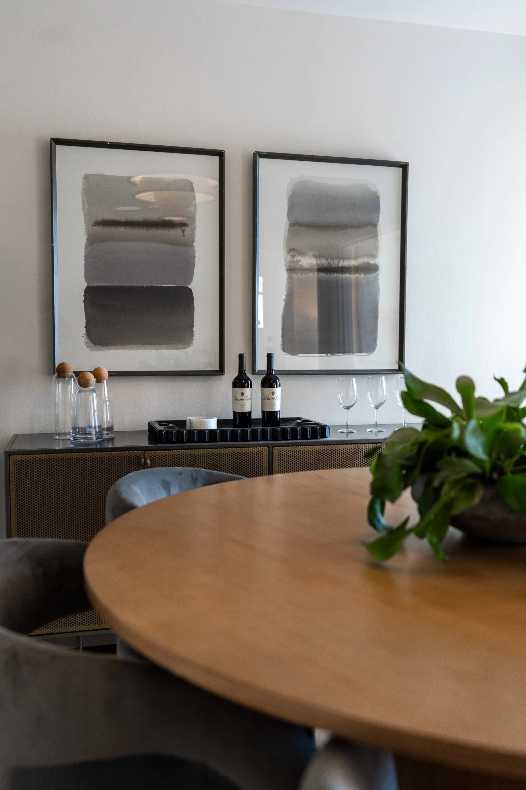 A dining area with a round wooden table in the foreground, a black sideboard with two bottles of wine, glasses, and framed abstract artworks on the wall, along with some decorative elements like bottles and a plant.