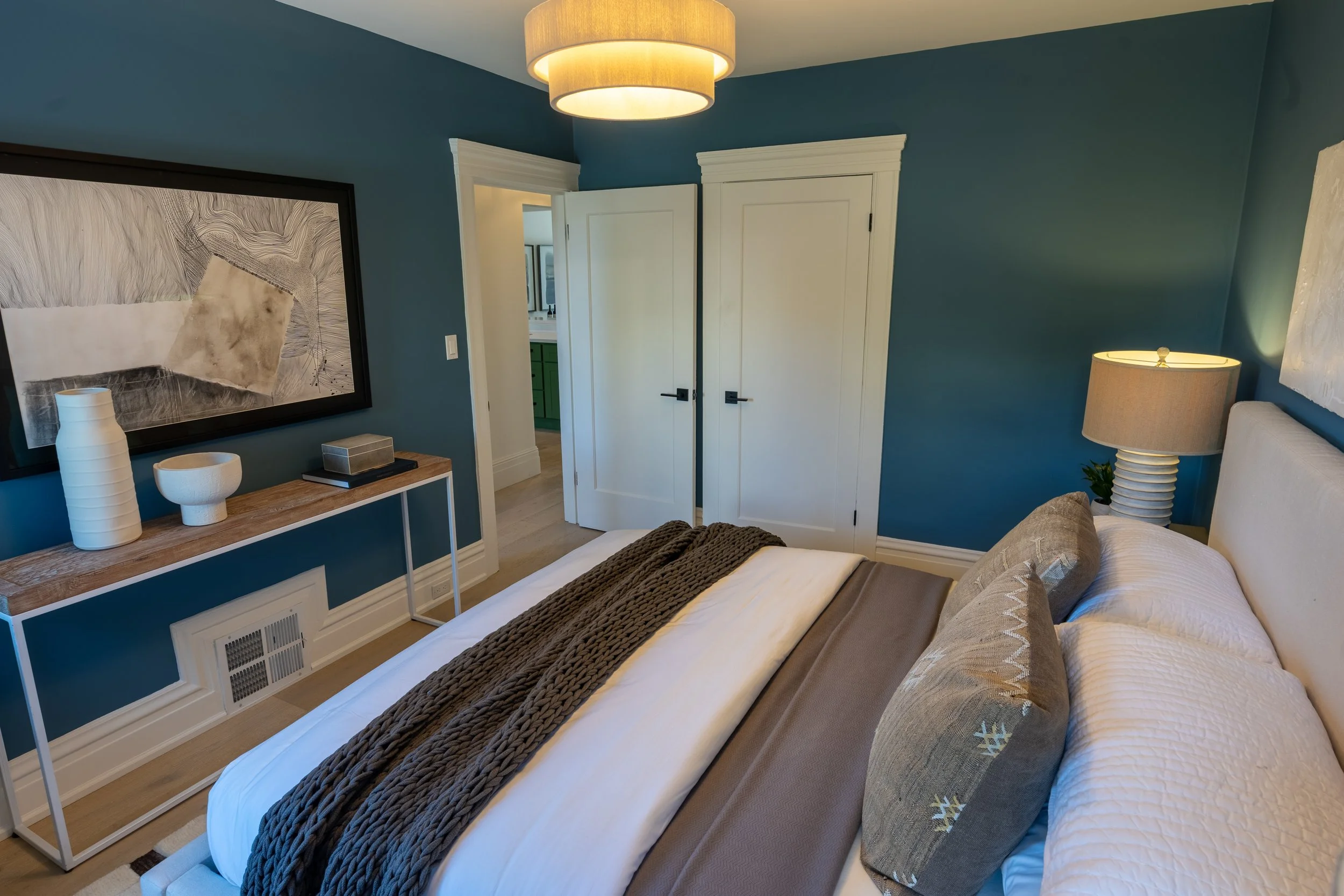 A bedroom with a blue wall, a bed with white bedding and gray pillows, a nightstand with a lamp, a large framed abstract artwork, a console table with decorative vases, and a ceiling light fixture.