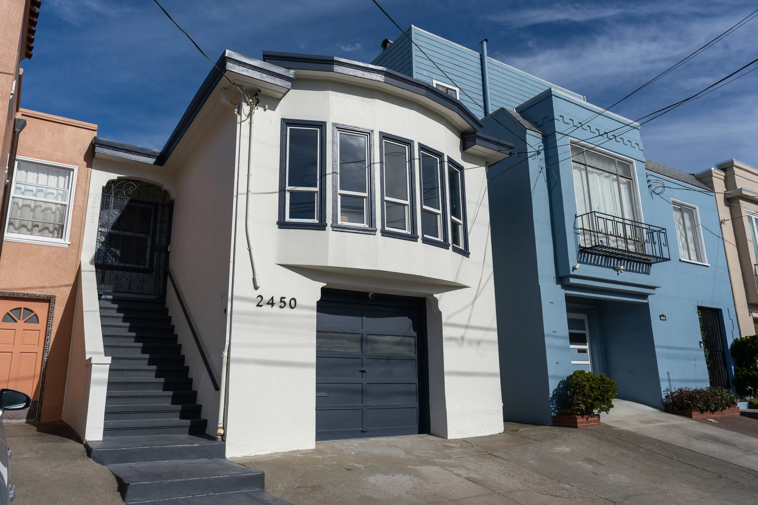 Row of colorful houses, one with a white facade and black trim, and another blue, with windows and balconies, under a clear blue sky with electrical wires overhead.