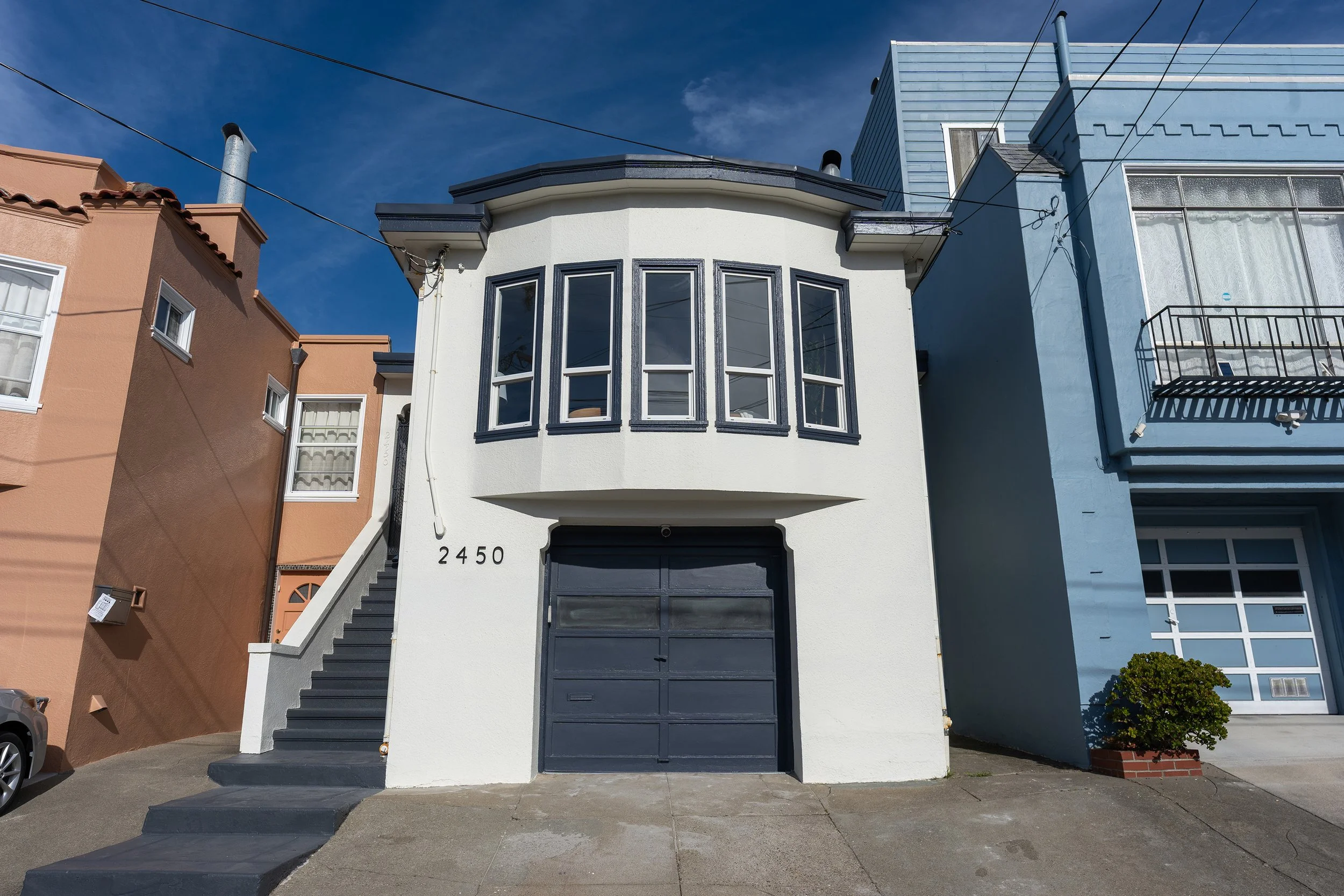 Front view of a white two-story house with black accents, a bay window, dark stairs leading to the entrance, and neighboring houses on either side under a blue sky.
