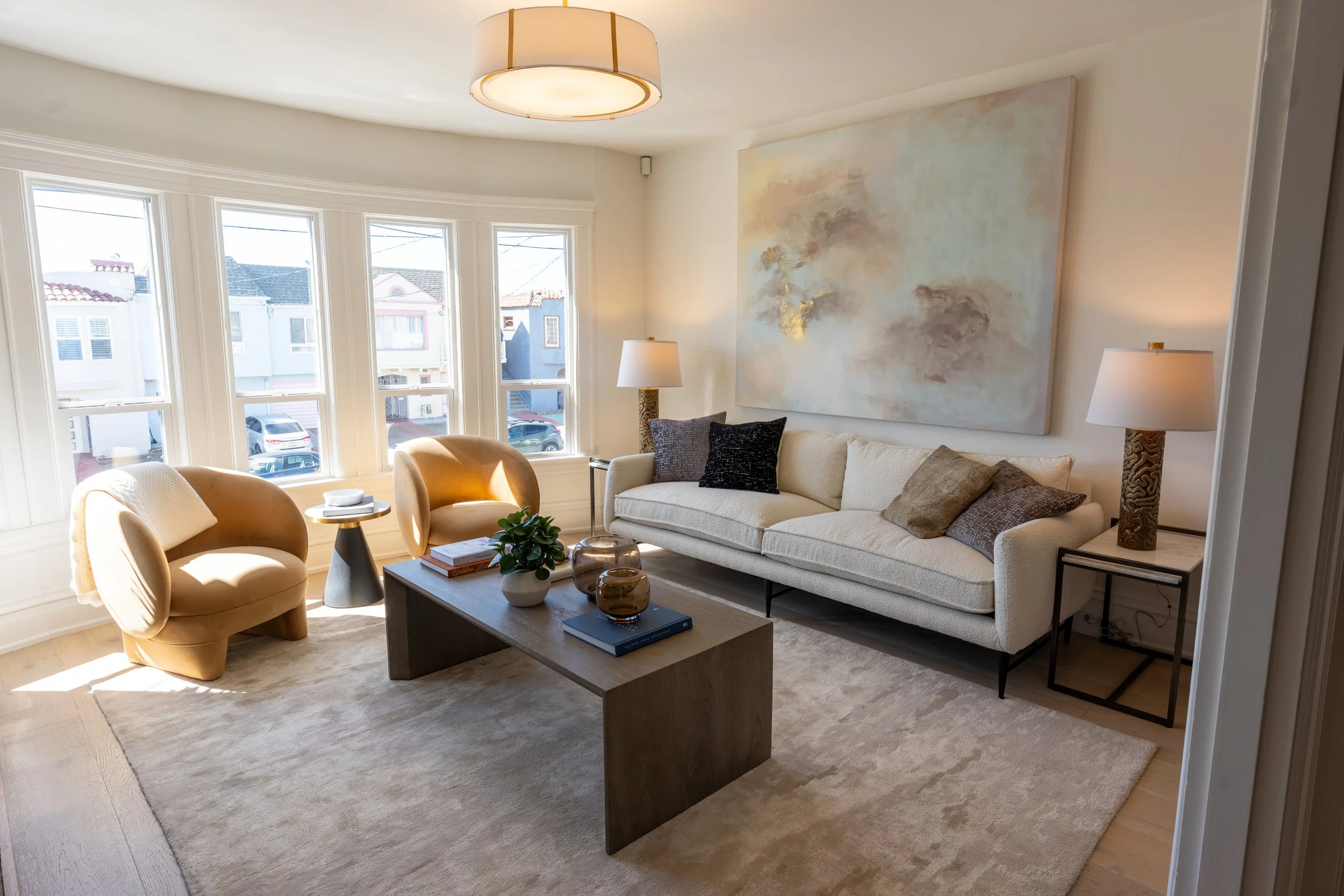 Living room with large windows, a beige sofa, a cream armchair, a wooden coffee table with decorative items, and artwork on the wall.