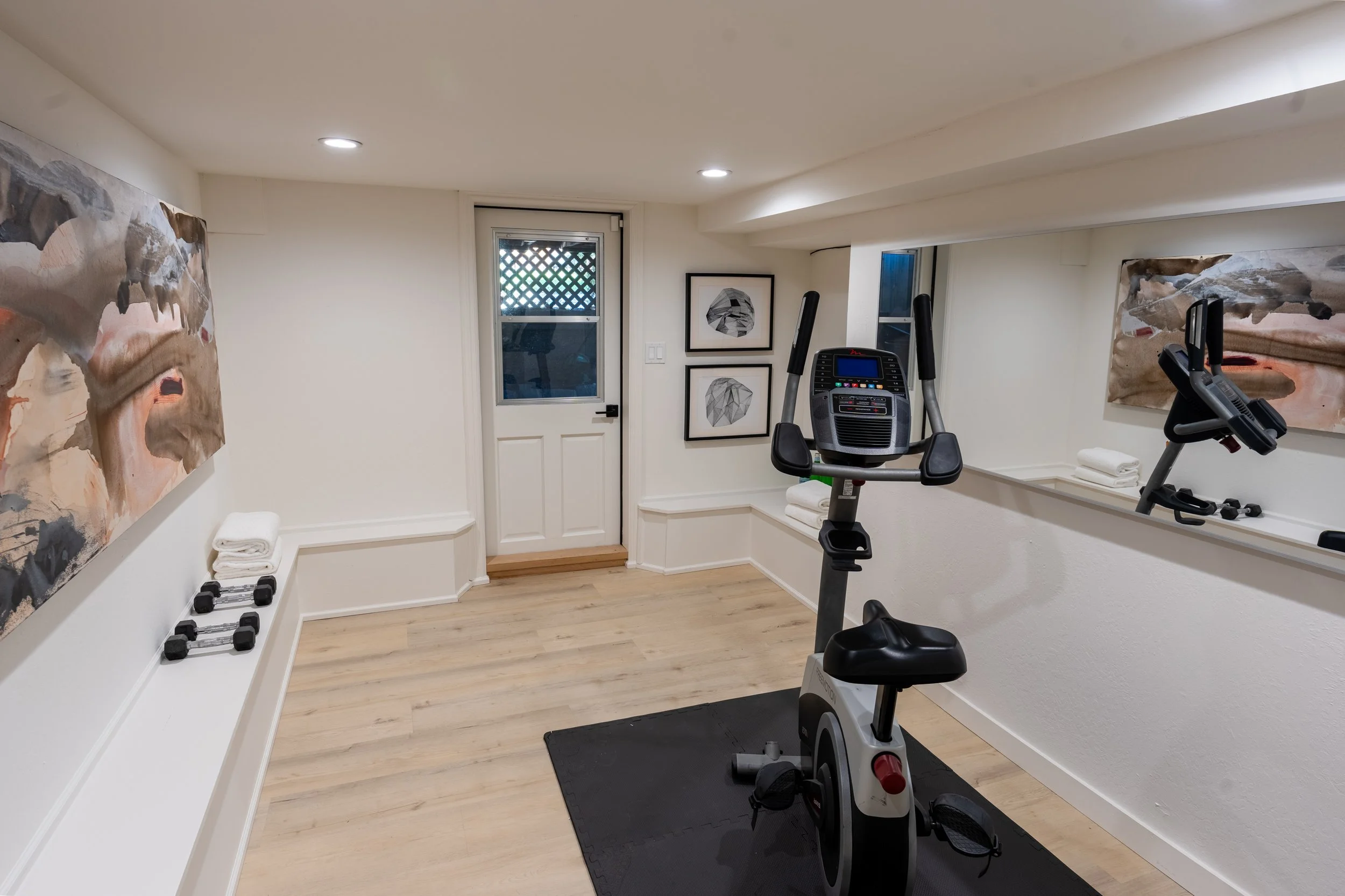 Home gym with exercise bike, elliptical machine, framed art on wall, dumbbells, and towels on shelf.
