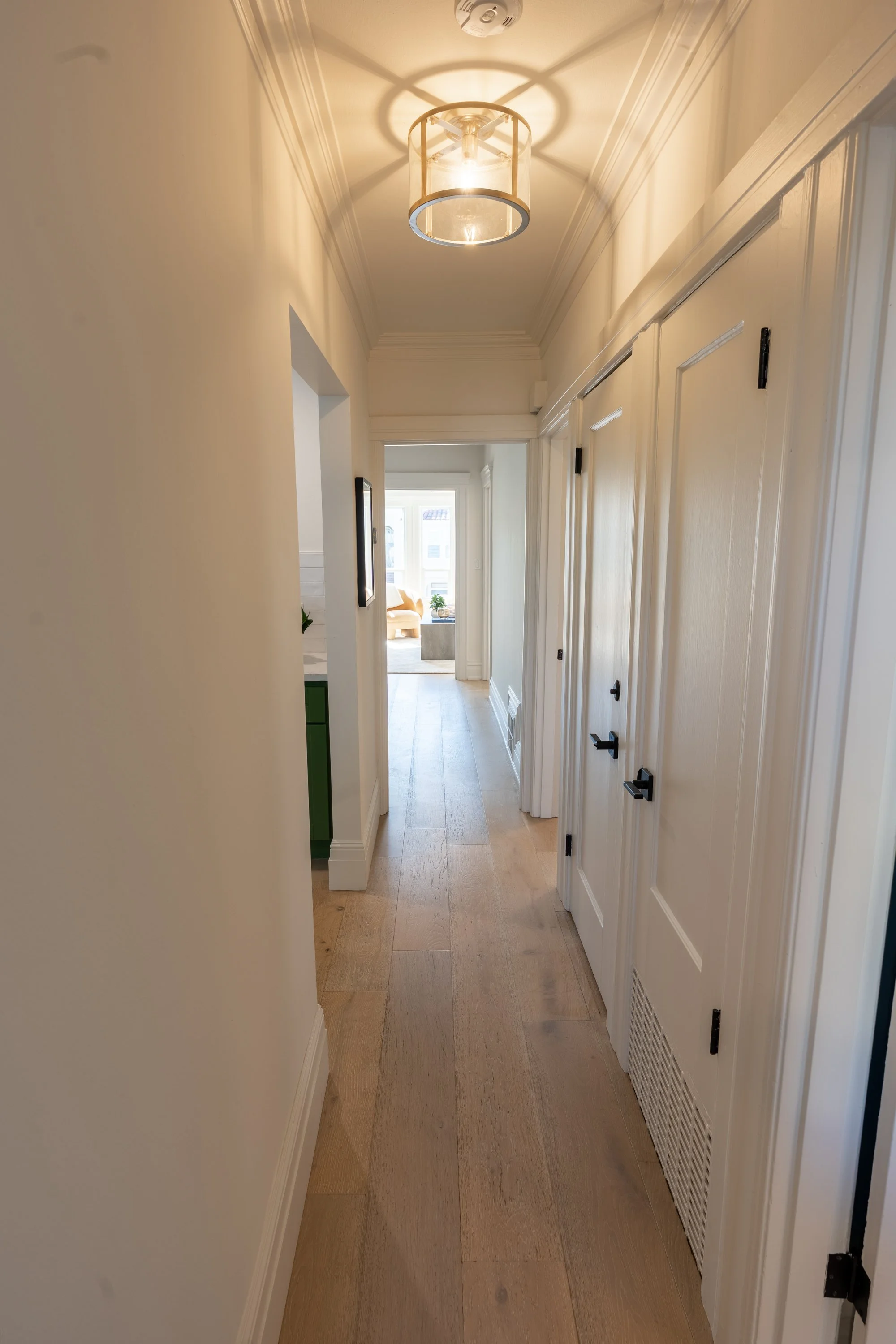 View of a narrow hallway with light-colored wooden flooring and white walls. A ceiling light fixture is visible, casting shadows on the ceiling. At the end of the hallway, a sunlit room with a window and a chair can be seen.