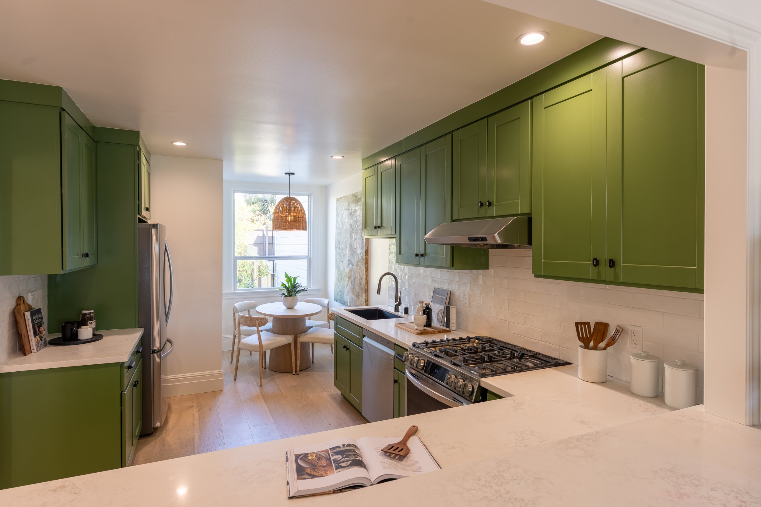 A modern kitchen with green cabinets, a white countertop, a stainless steel stove, and a window with a view of trees. There is a small round dining table with four chairs and a hanging wicker lamp.