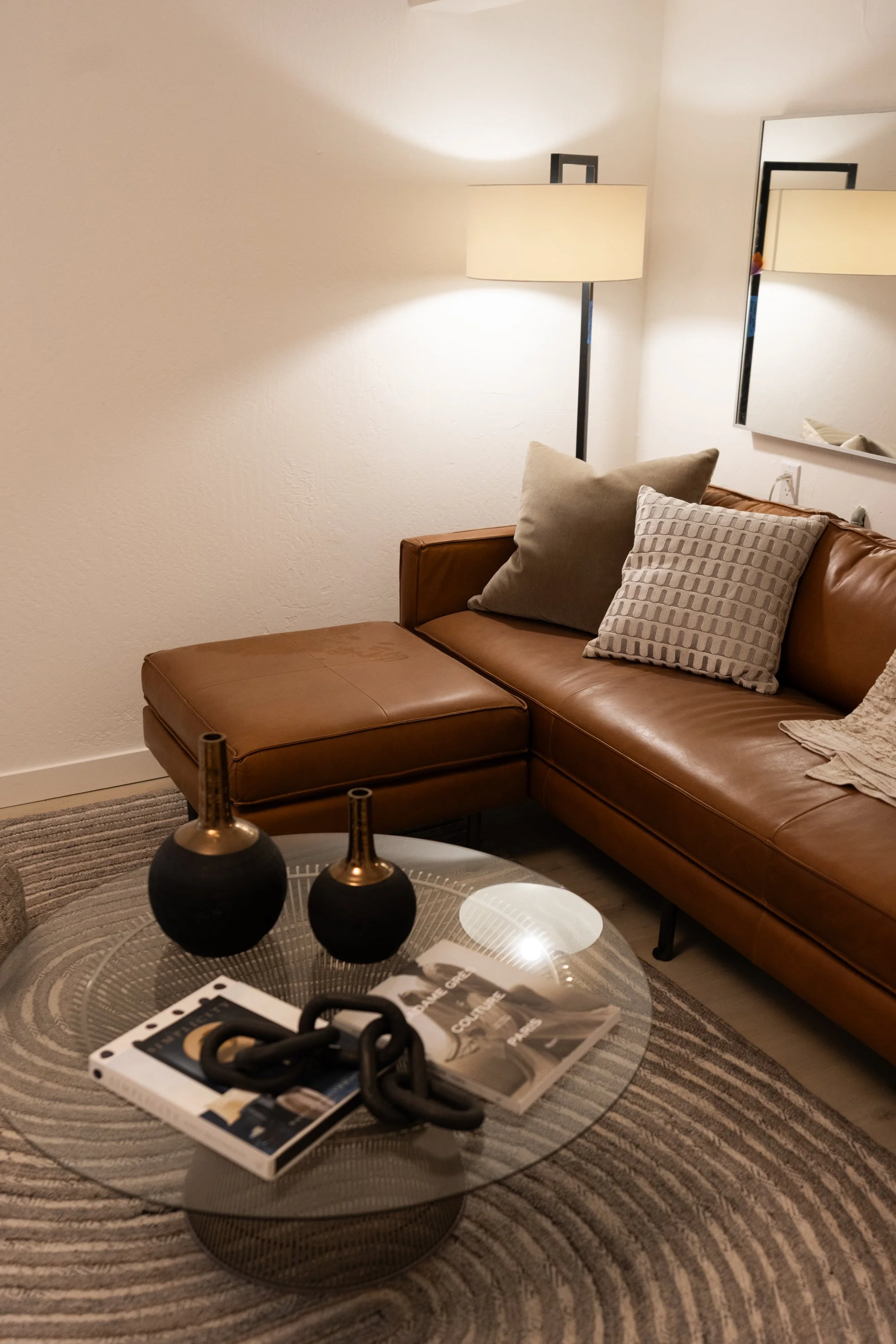 Living room with a brown leather sofa, beige pillows, a floor lamp, mirror on the wall, a glass coffee table with black decorative objects, magazines, and a rug.