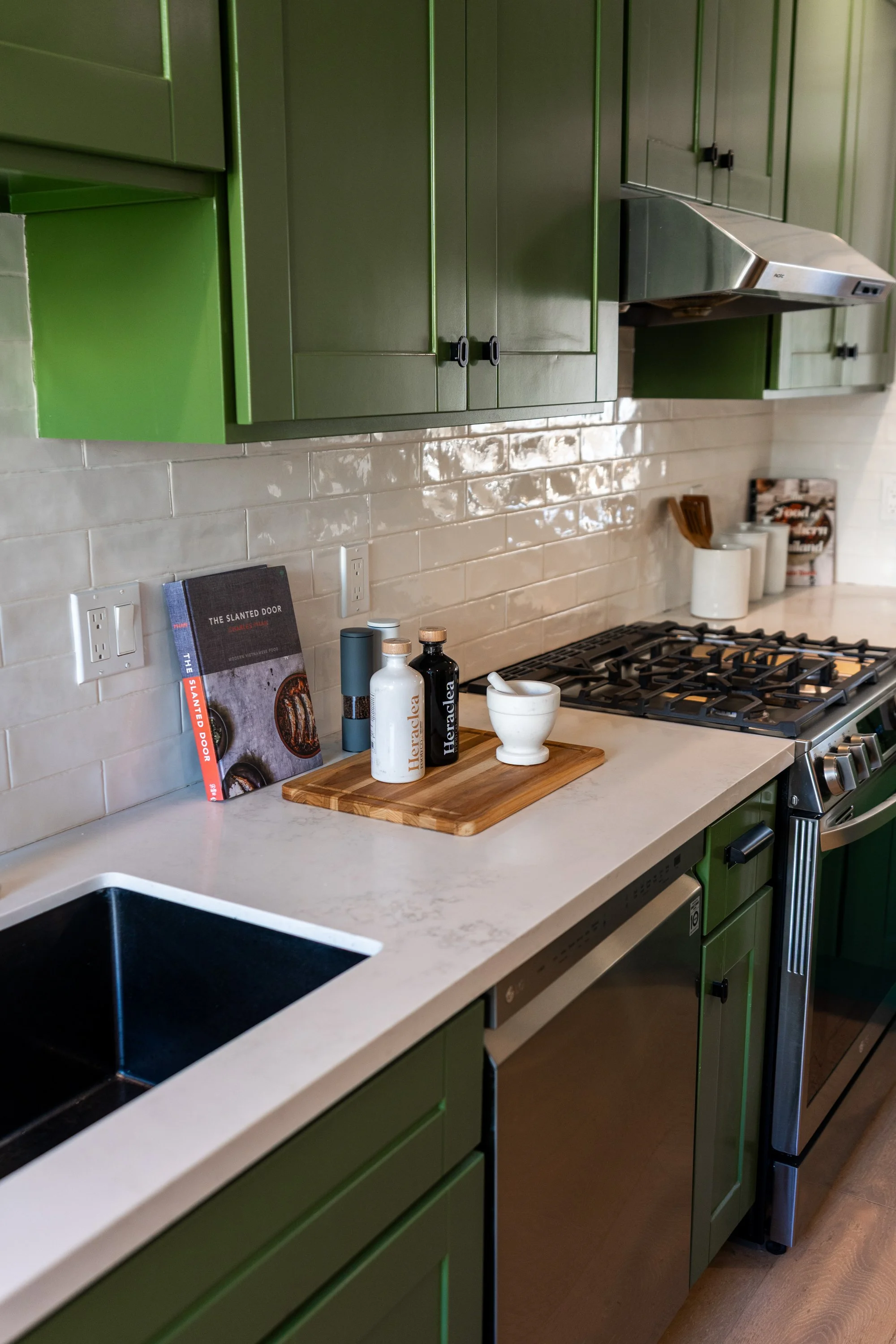Green kitchen cabinets with a white countertop, a black sink, and cooking appliances. Kitchen accessories include books, bottles, and a wooden cutting board.