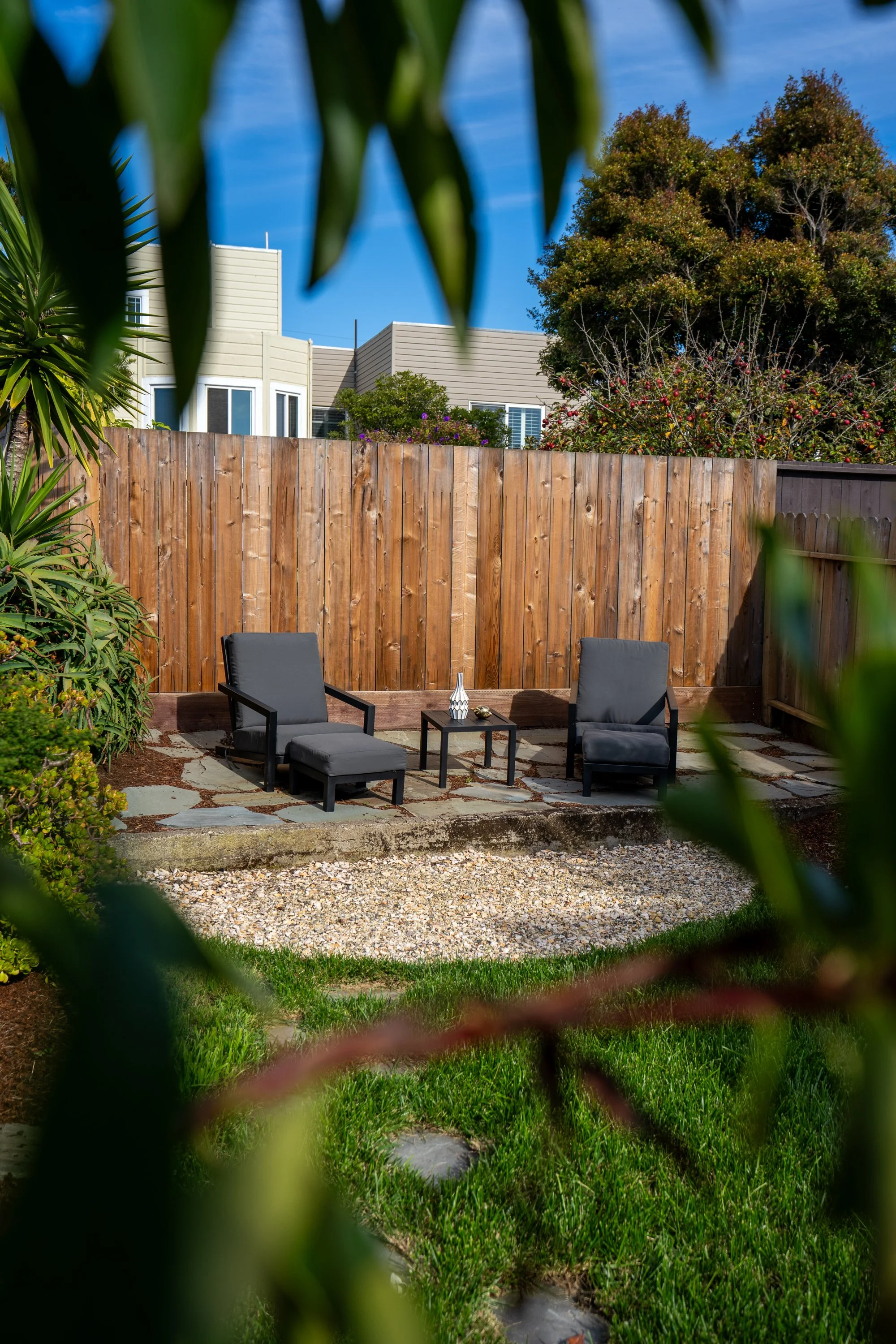 A backyard patio area with two black chairs and a small table, surrounded by a wooden fence and lush green plants, with residential houses and trees in the background under a clear blue sky.