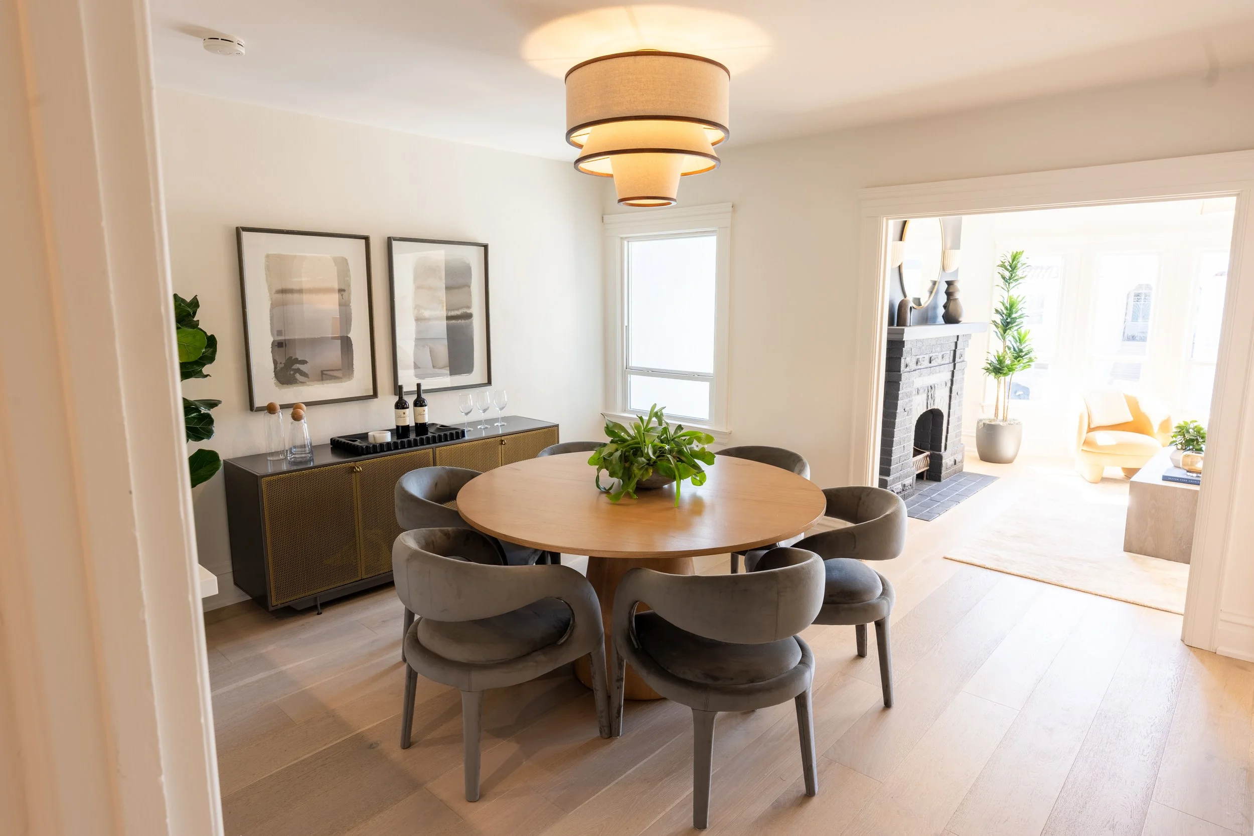 A dining room with a round wooden table surrounded by six gray upholstered chairs, a black sideboard with wine bottles and glasses, modern abstract artwork on the wall, and a view into a living room with a gray brick fireplace, a yellow armchair, and