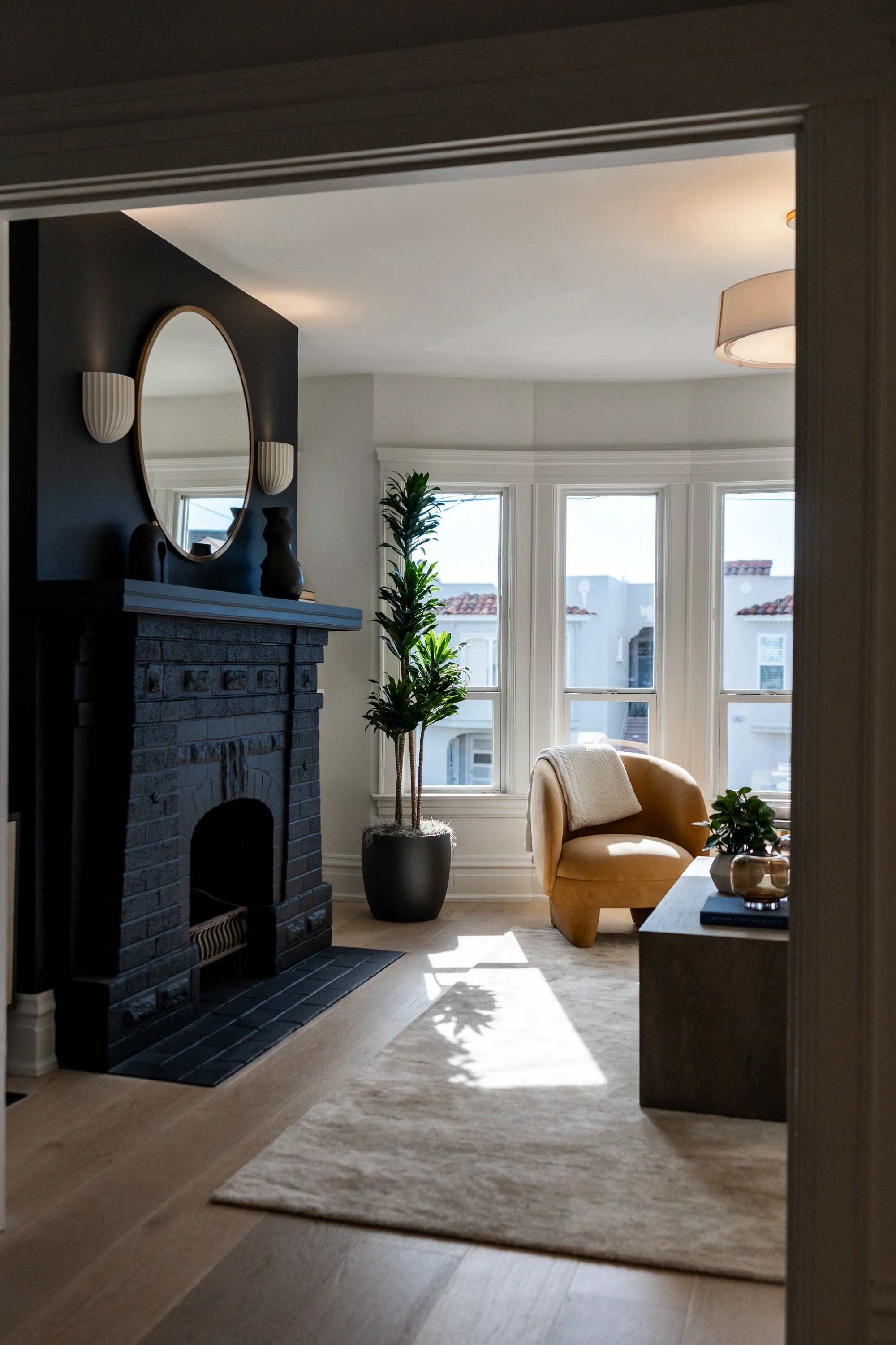 A cozy living room with a black brick fireplace, a mirror above it, and decorative vases. Large bay windows let in natural light, illuminating a tan armchair, a potted plant, a side table with books, and a neutral-colored rug on light wood flooring.
