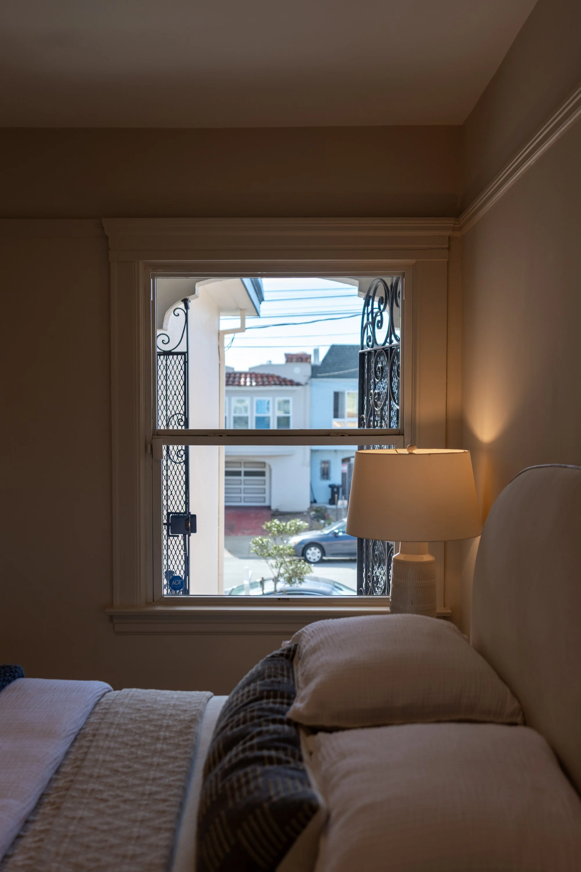 A view from inside a bedroom looking out the window toward a street with parked cars and neighboring houses. The bed with white and plaid bedding is partially visible in the foreground, and a nightstand with a lamp is next to the bed.
