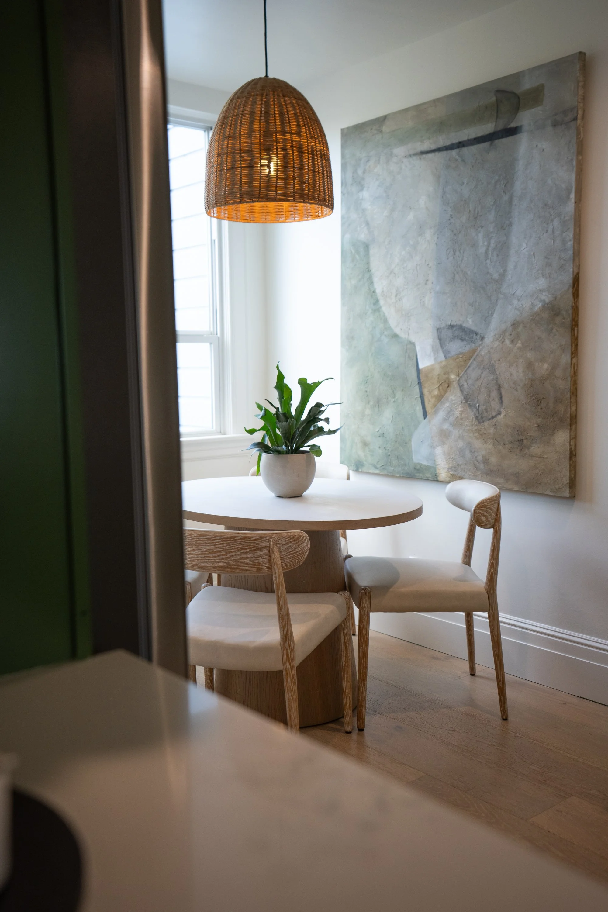 A dining table with four wooden chairs and a potted green plant on top, located in front of a large abstract watercolor artwork on a white wall. Hanging above is a woven rattan pendant light near a window.
