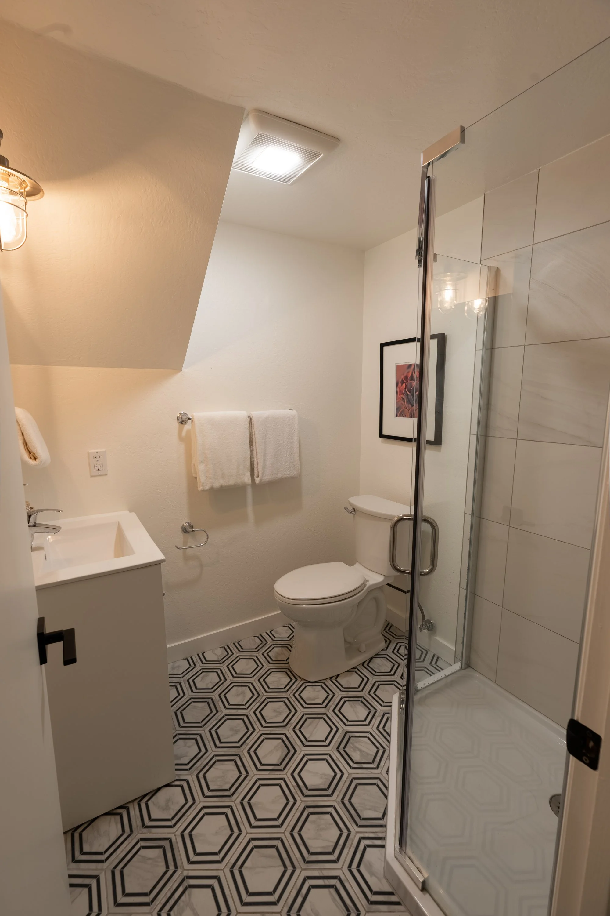 A small modern bathroom with a black and white geometric patterned floor, white walls, a toilet, a sink vanity, two white towels on a towel bar, framed artwork on the wall, a glass shower enclosure, and a ceiling light.