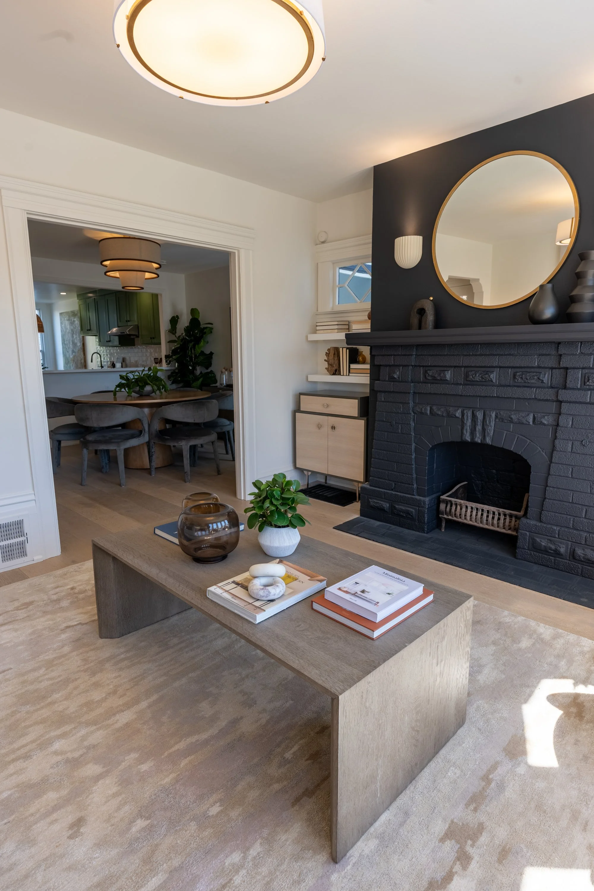 Living room with black brick fireplace, round mirror, and minimalist decor, open to dining area with green cabinets and round table.