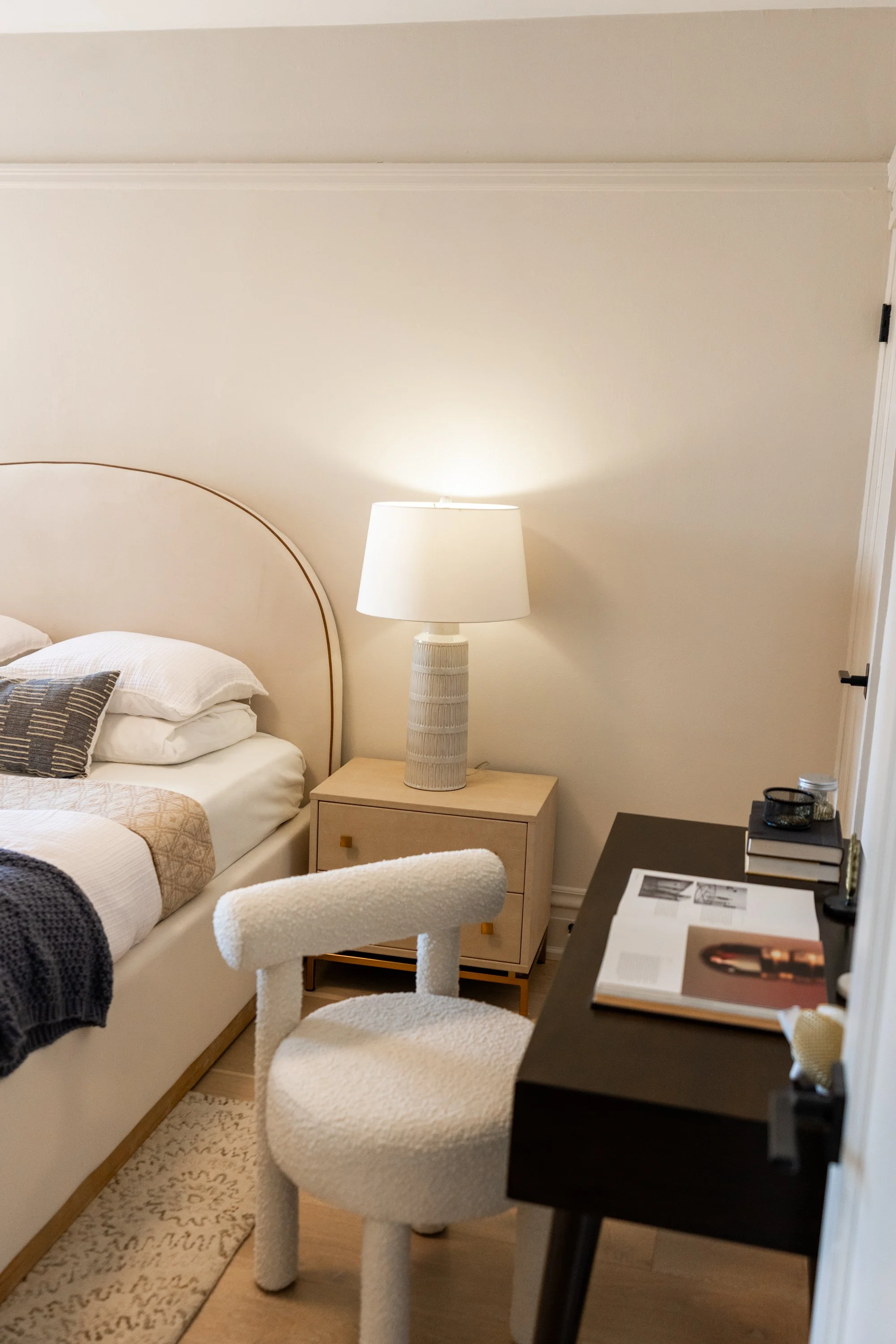 A bedroom scene with a bed, nightstand, table lamp, and a black desk with books and papers, and a white chair with textured upholstery.