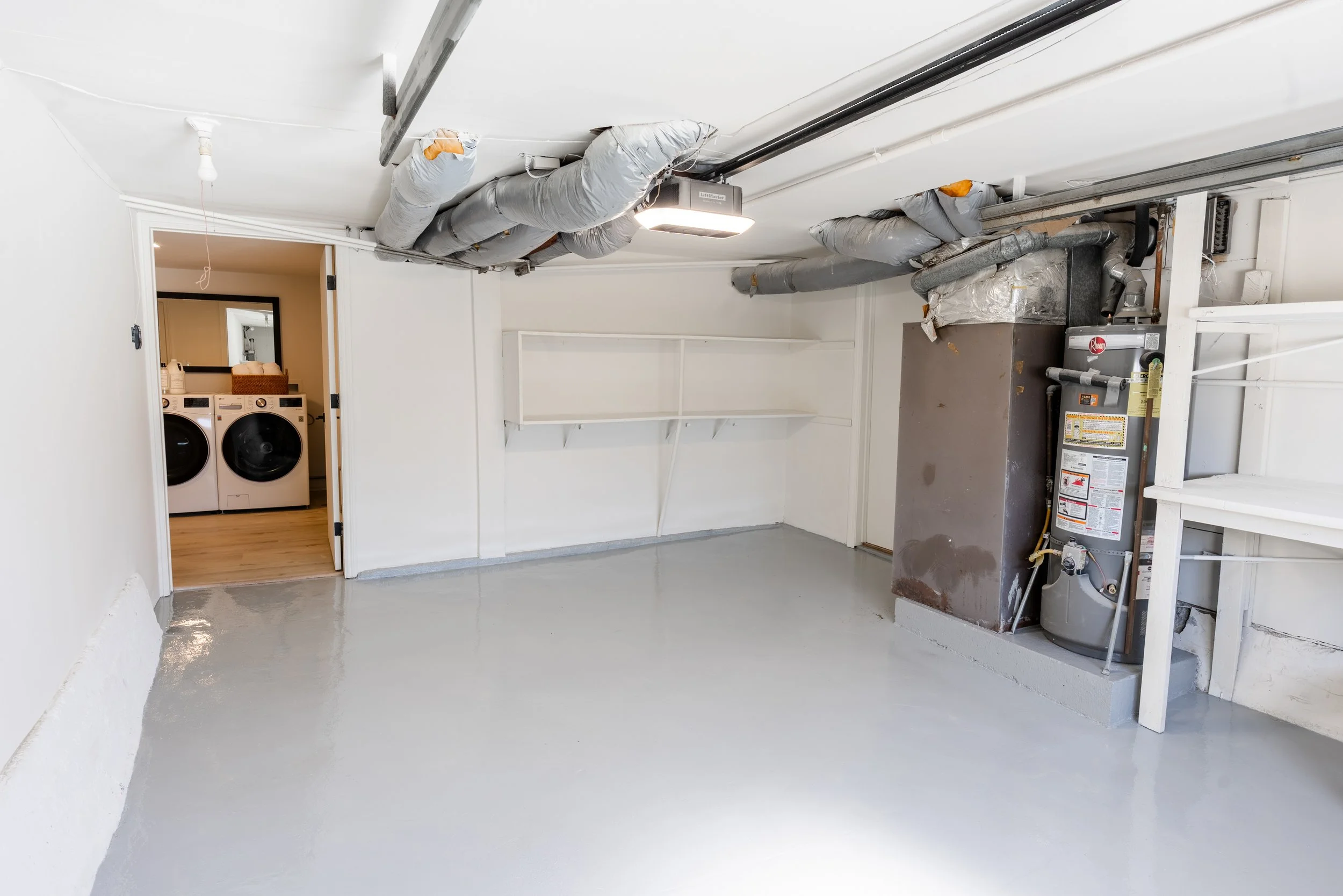 Empty laundry room with white walls, a washing machine and dryer visible through an open door, and HVAC ductwork on the ceiling.