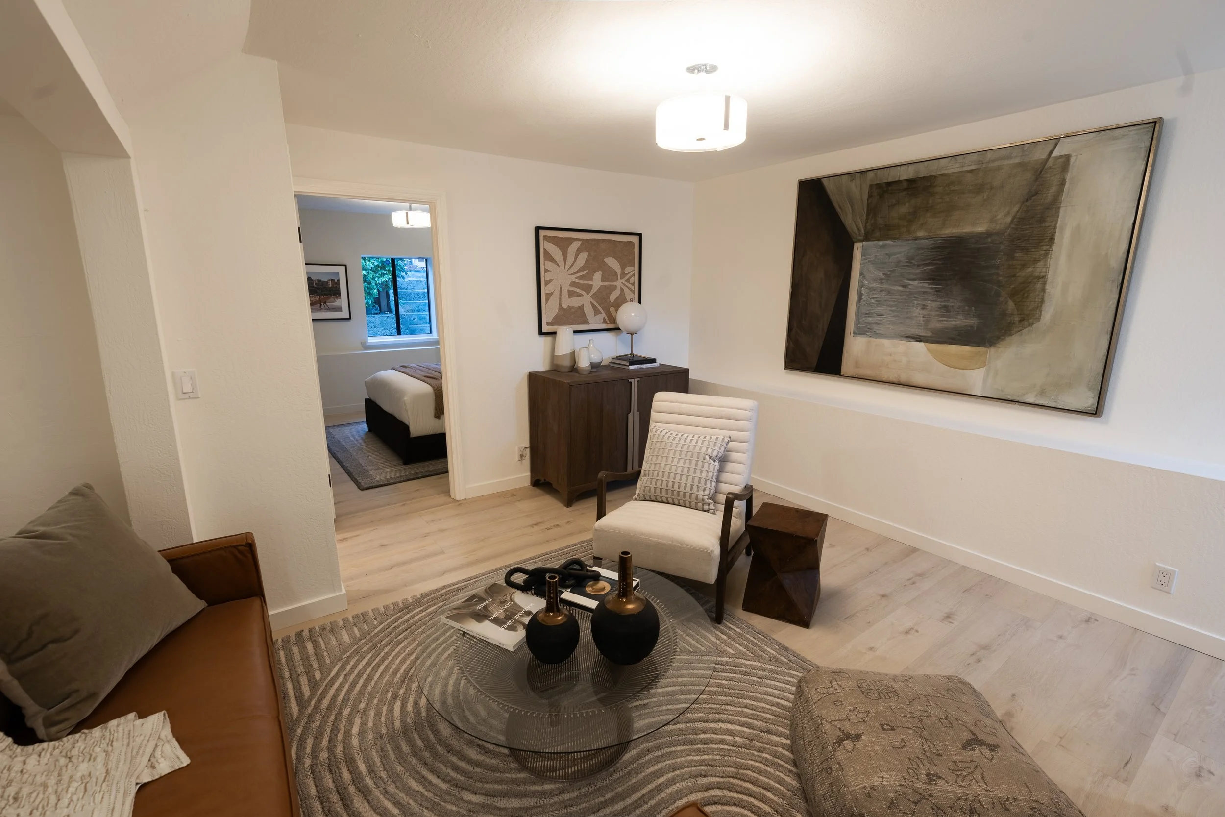 Living room with a tan sofa, a white armchair, artwork on the walls, a wooden cabinet, and a coffee table with decorative vases and black objects.