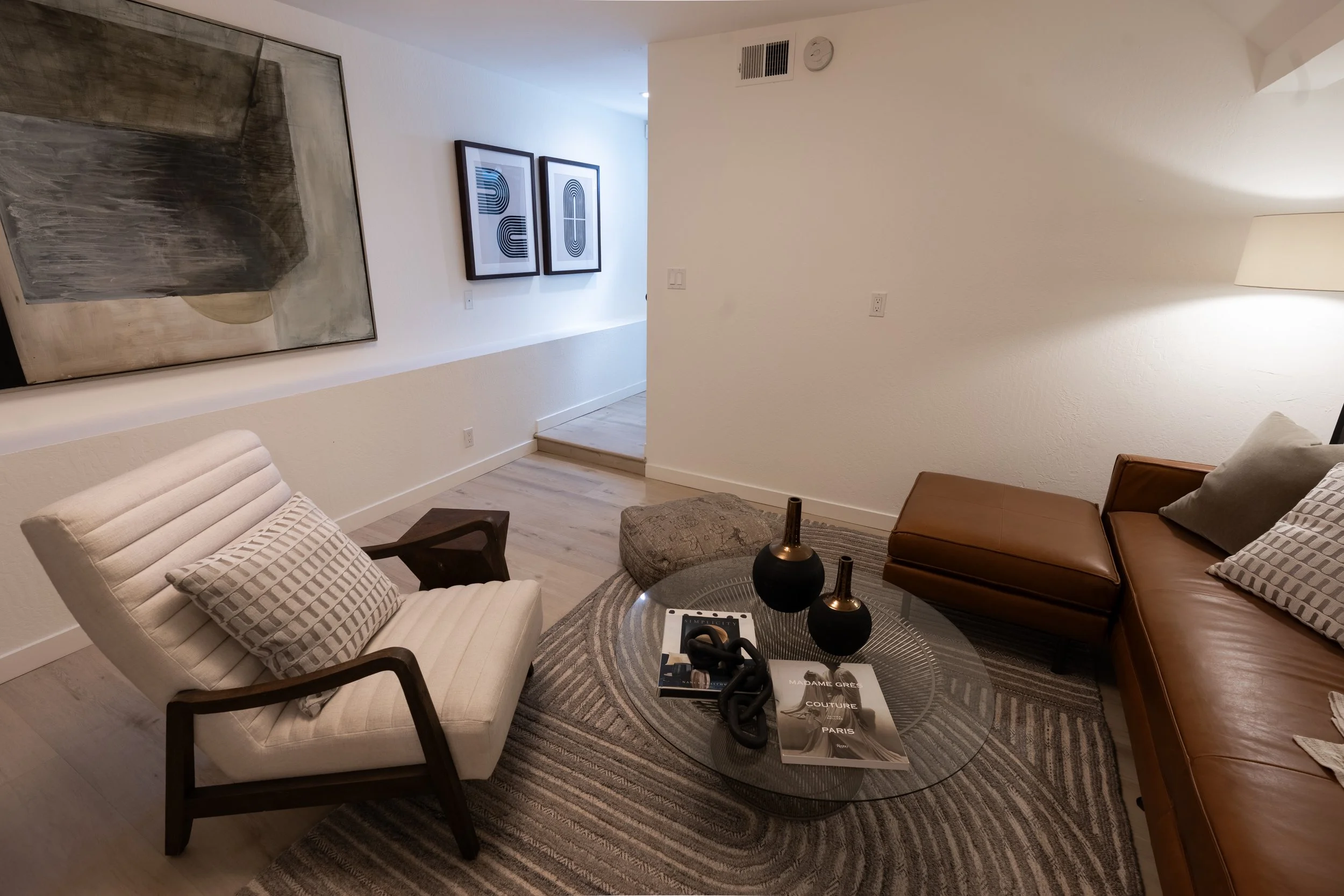 Living room with beige and brown furniture, including an upholstered chair with a striped pillow, a leather sofa with pillows, a round glass table with decorative vases, and wall art featuring abstract black and white designs. The room has light-colo