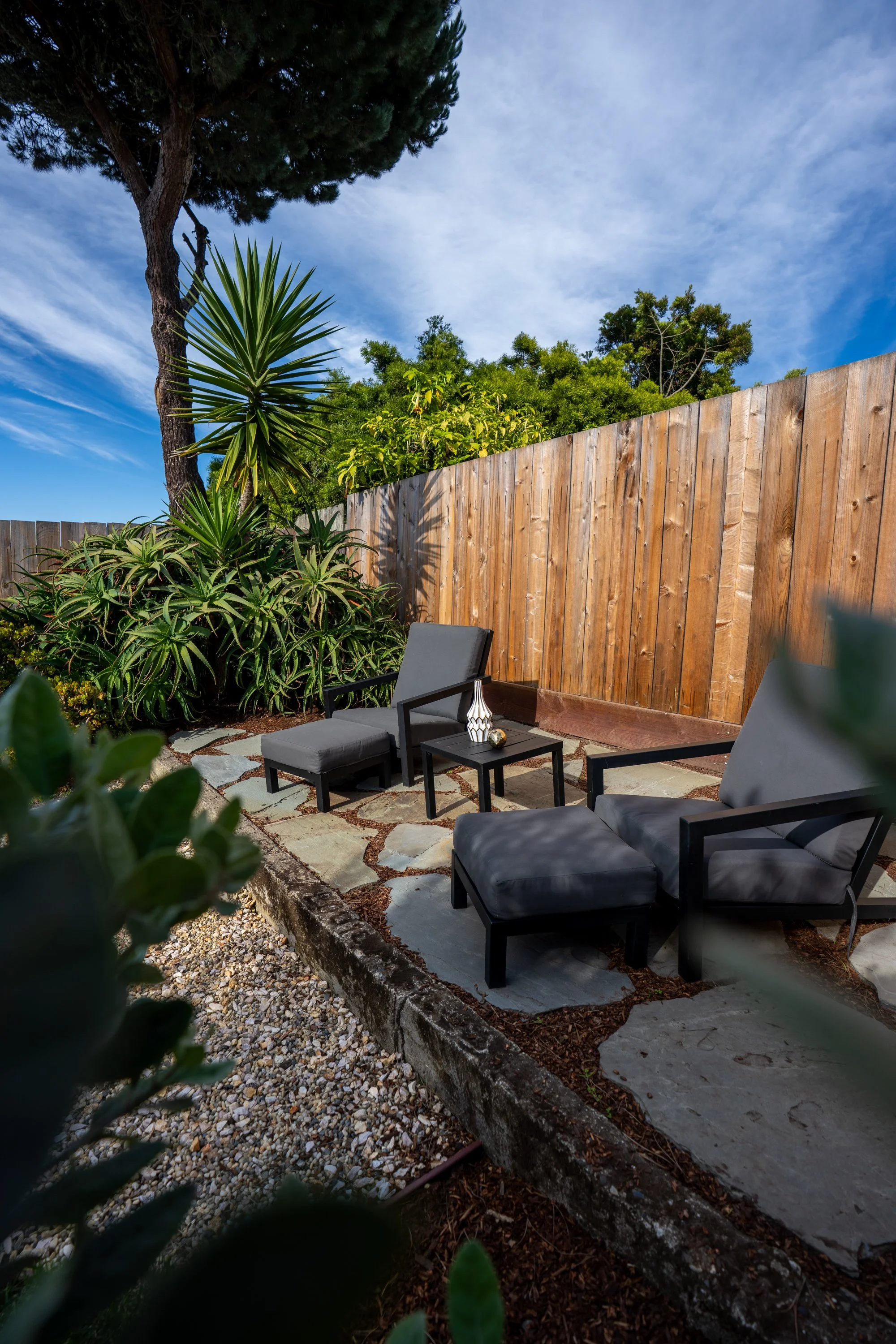A small outdoor patio area with black chairs and a small black table with decorative objects, surrounded by green bushes and trees, wooden fence in the background, and a partly cloudy blue sky overhead.