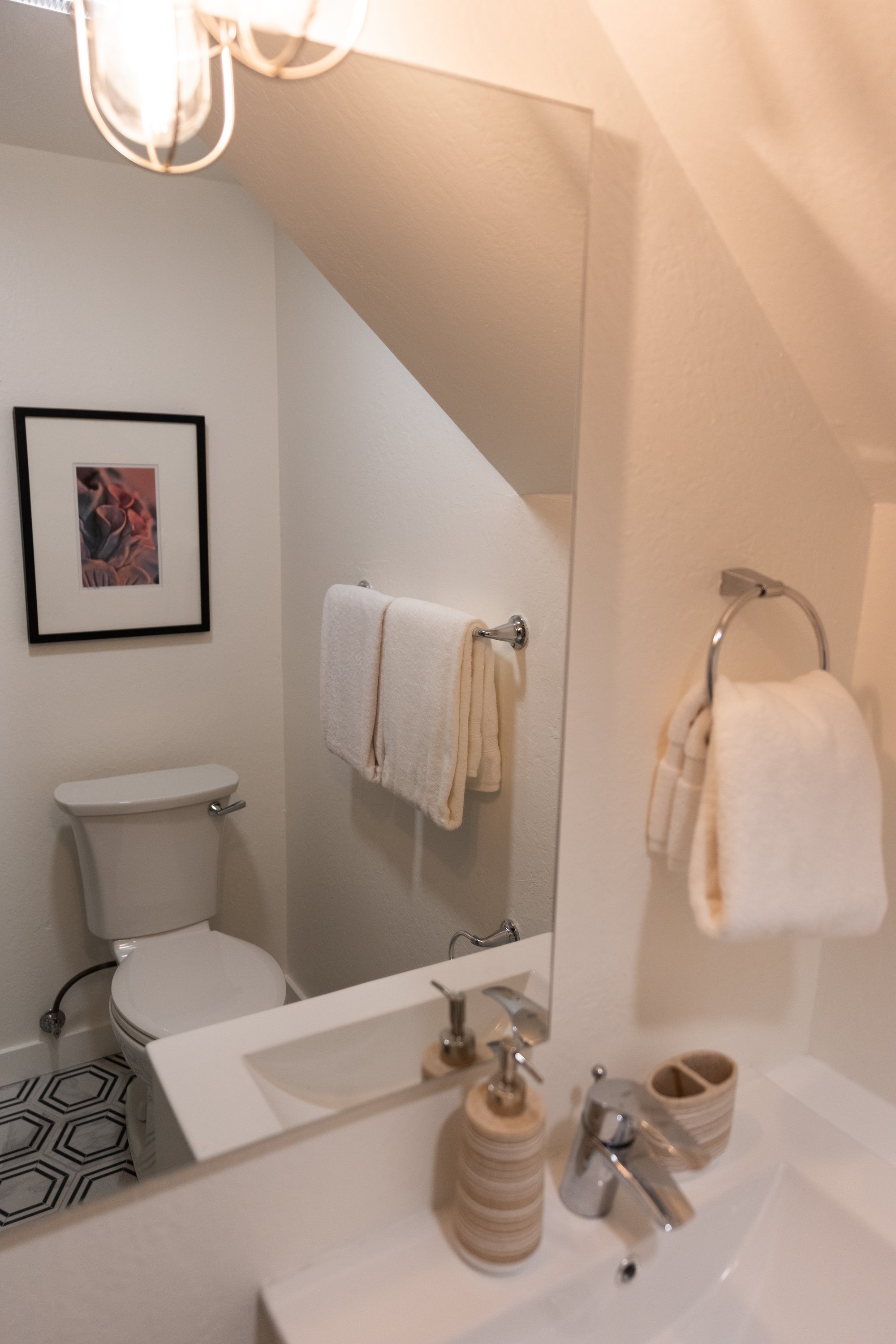 A small bathroom with a white toilet, a mirror, a white sink, and beige towels hanging on a silver towel rack. The walls are white with a framed abstract art piece. The floor has black and white hexagonal tiles, and there is a bathroom countertop wit