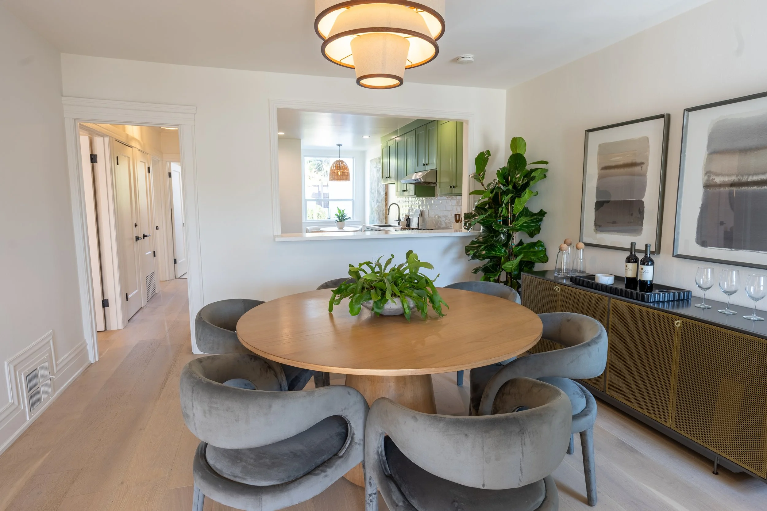 Modern dining room with a round wooden table surrounded by gray velvet chairs. There are green plants on and near the table, and framed abstract art on the wall. In the background, an open kitchen with green cabinets and a window letting in natural l