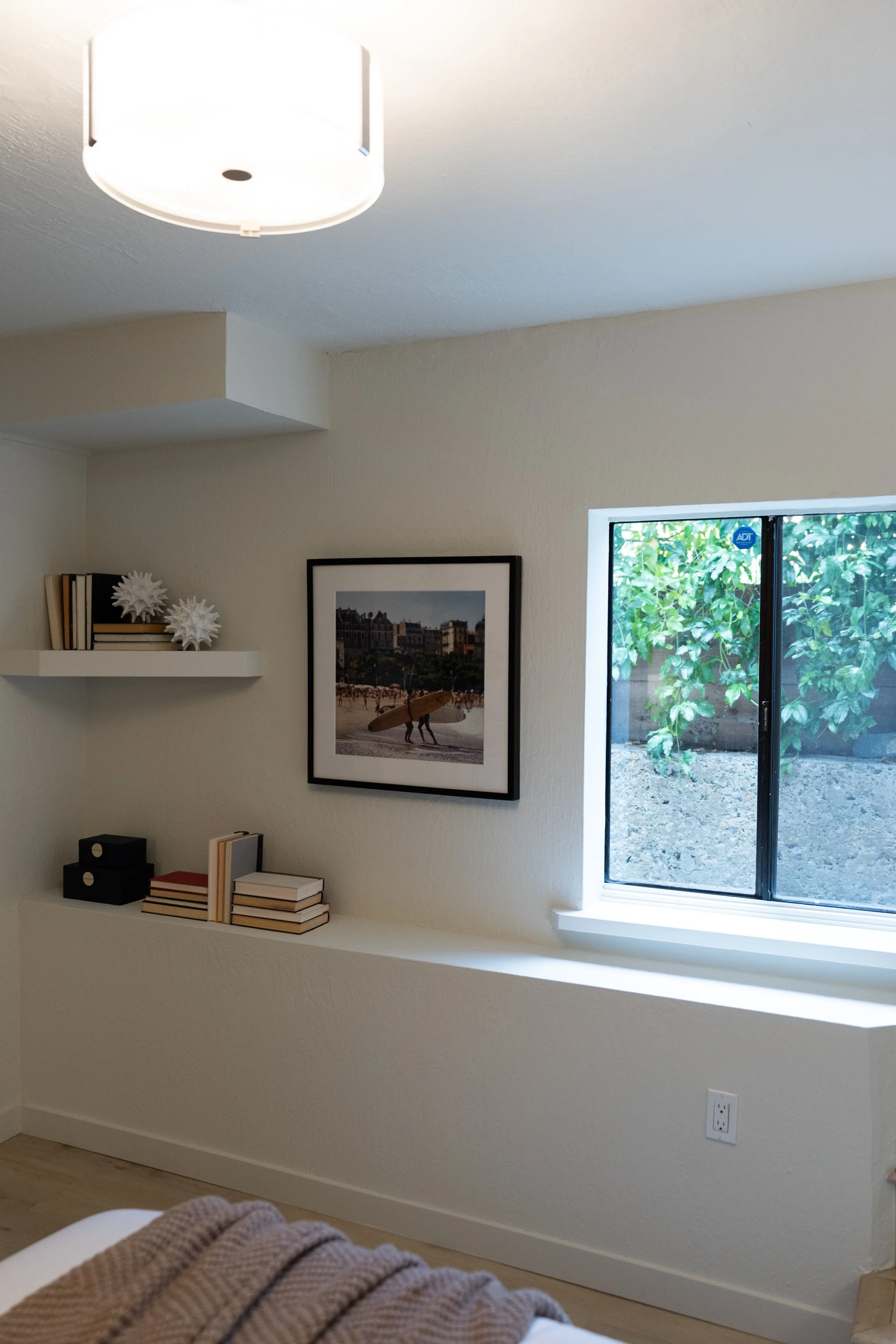 A corner of a room with beige walls, a window with green foliage outside, a framed beach scene photograph, white floating shelves with decorative objects, and a dresser with books. Part of a bed with a brown and beige striped blanket is visible in th