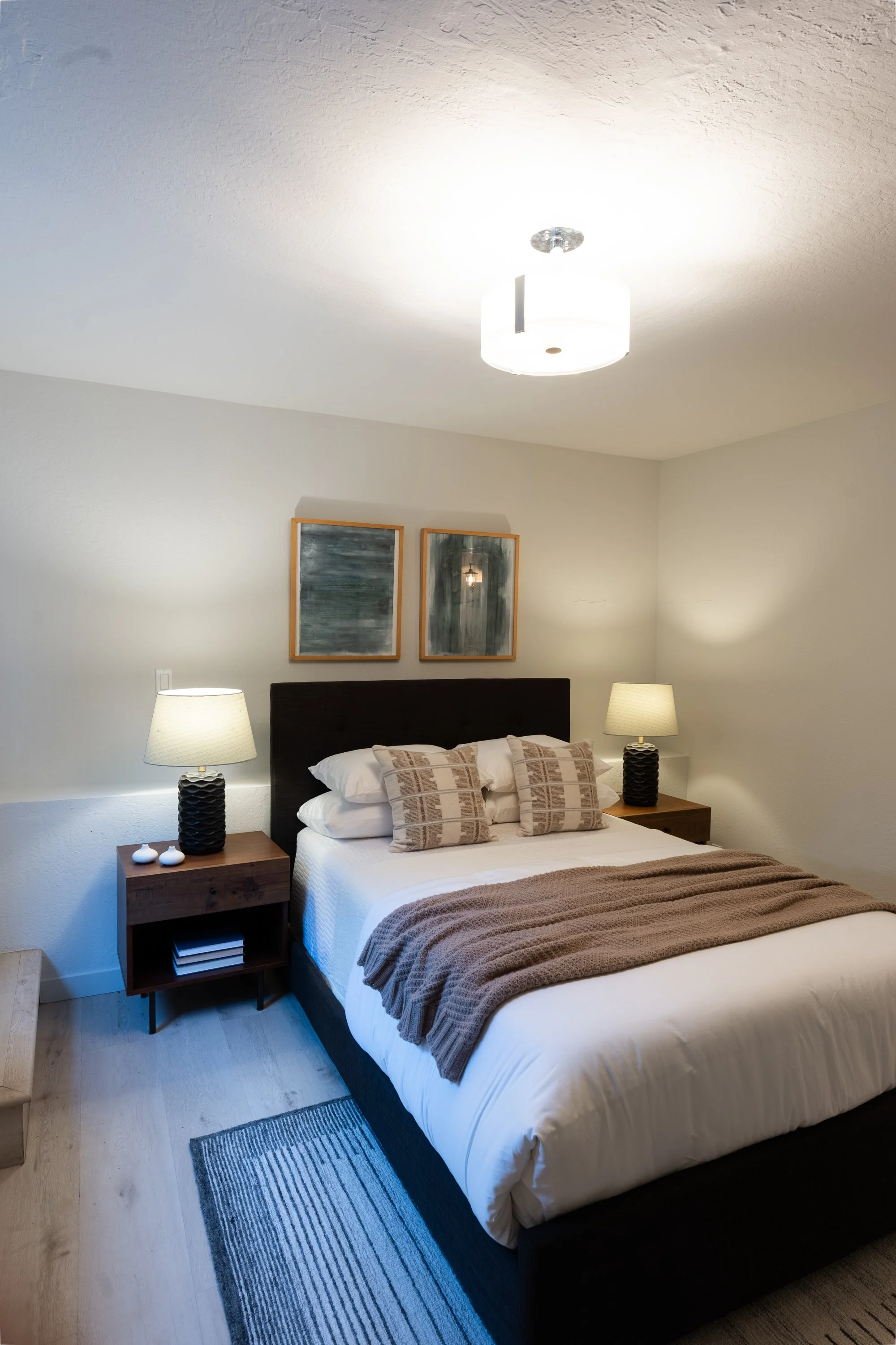 A bedroom with a bed covered in white sheets and a brown blanket at the foot. There are six beige pillows on the bed. Two wooden nightstands with lamps are on either side of the bed. The lamps have black, textured bases and white shades. Two framed a