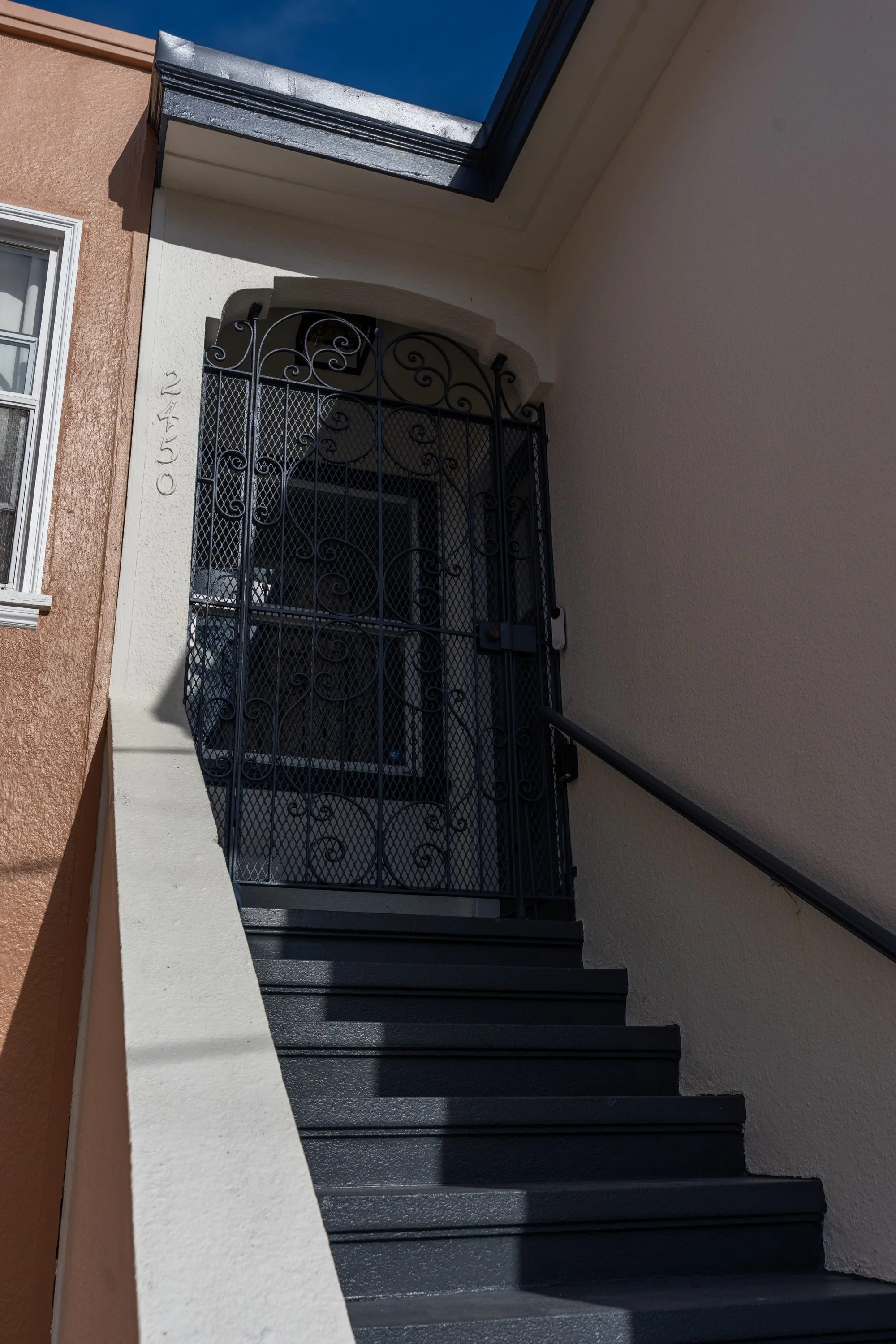 Exterior stairs leading to an entrance with a decorative black wrought iron gate, part of a building with beige and white walls, and a black staircase handrail, under a clear blue sky.